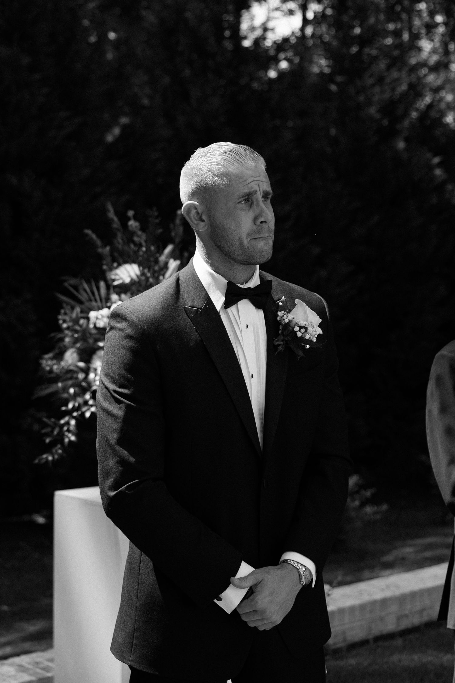 Groom in black tuxedo with boutonniere stands at altar during outdoor wedding ceremony, black and white photo.