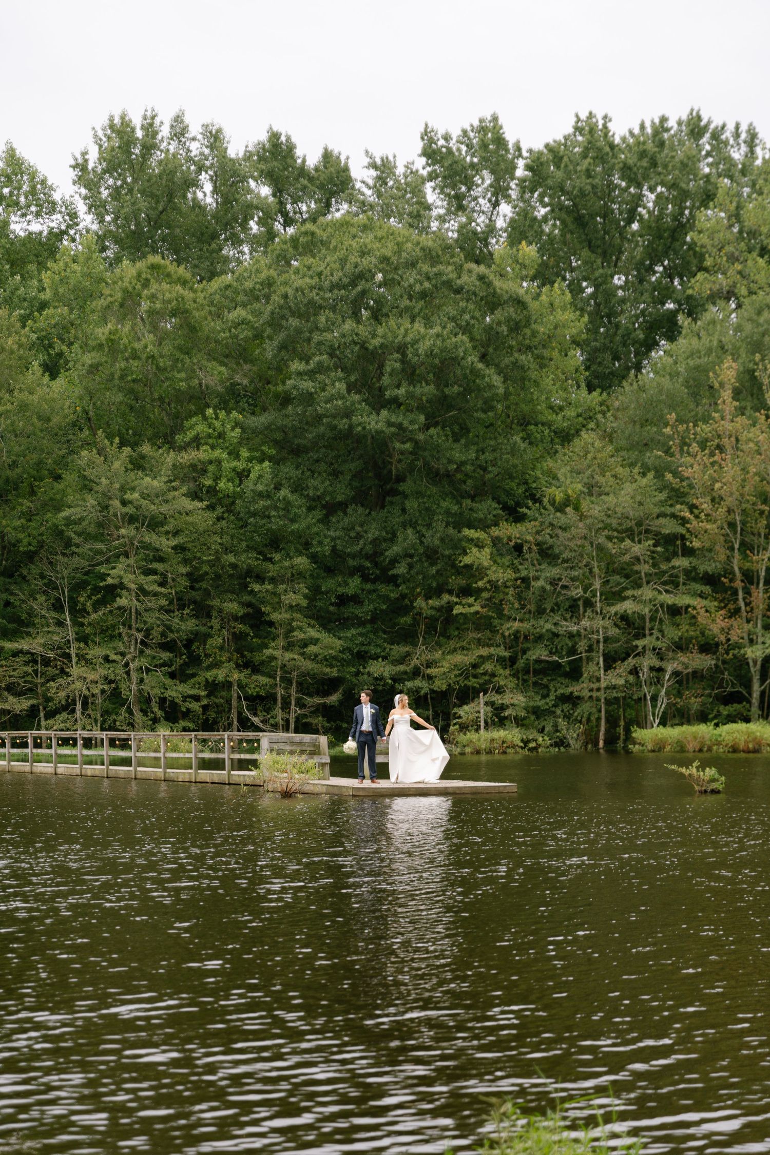 Bride and groom stand on a serene lakeside dock surrounded by lush green trees on their wedding day.