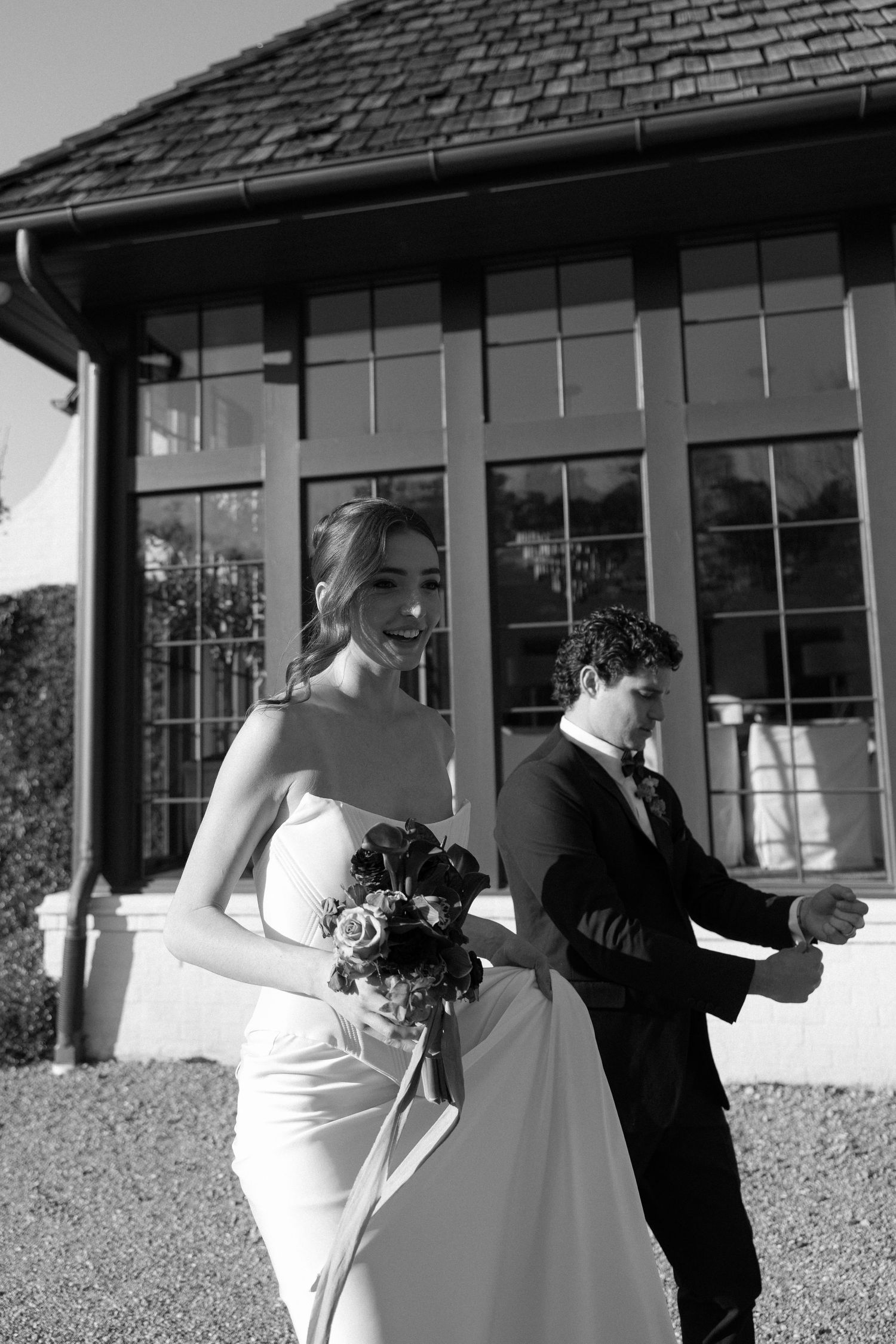 Black and white photo of a smiling bride holding a bouquet and groom in a suit outside an elegant building.