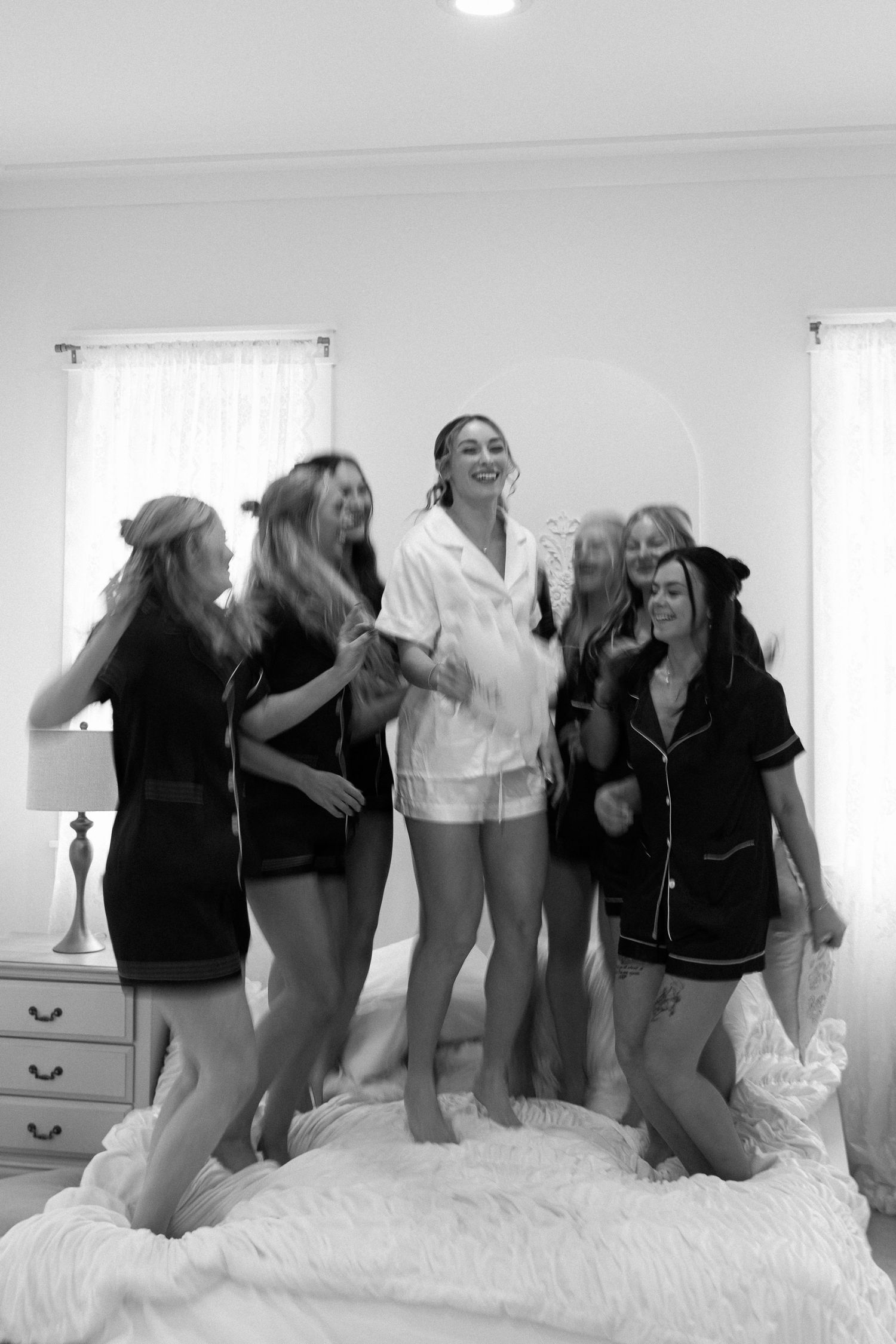 Bridesmaids in black pajamas jumping on bed with bride in white pajamas during bridal party getting ready.