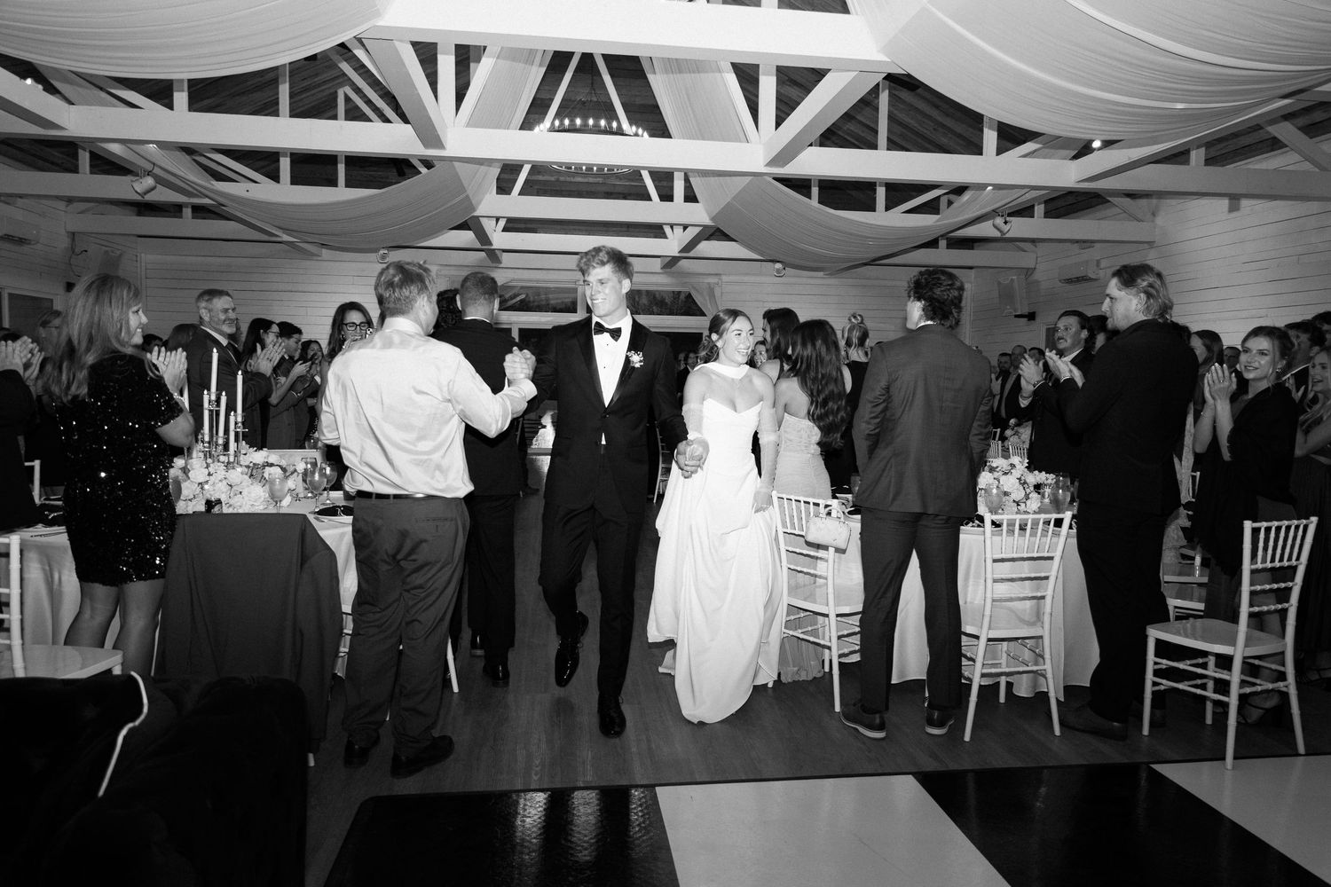Black and white photo of bride and groom making grand entrance at elegant wedding reception with guests applauding.