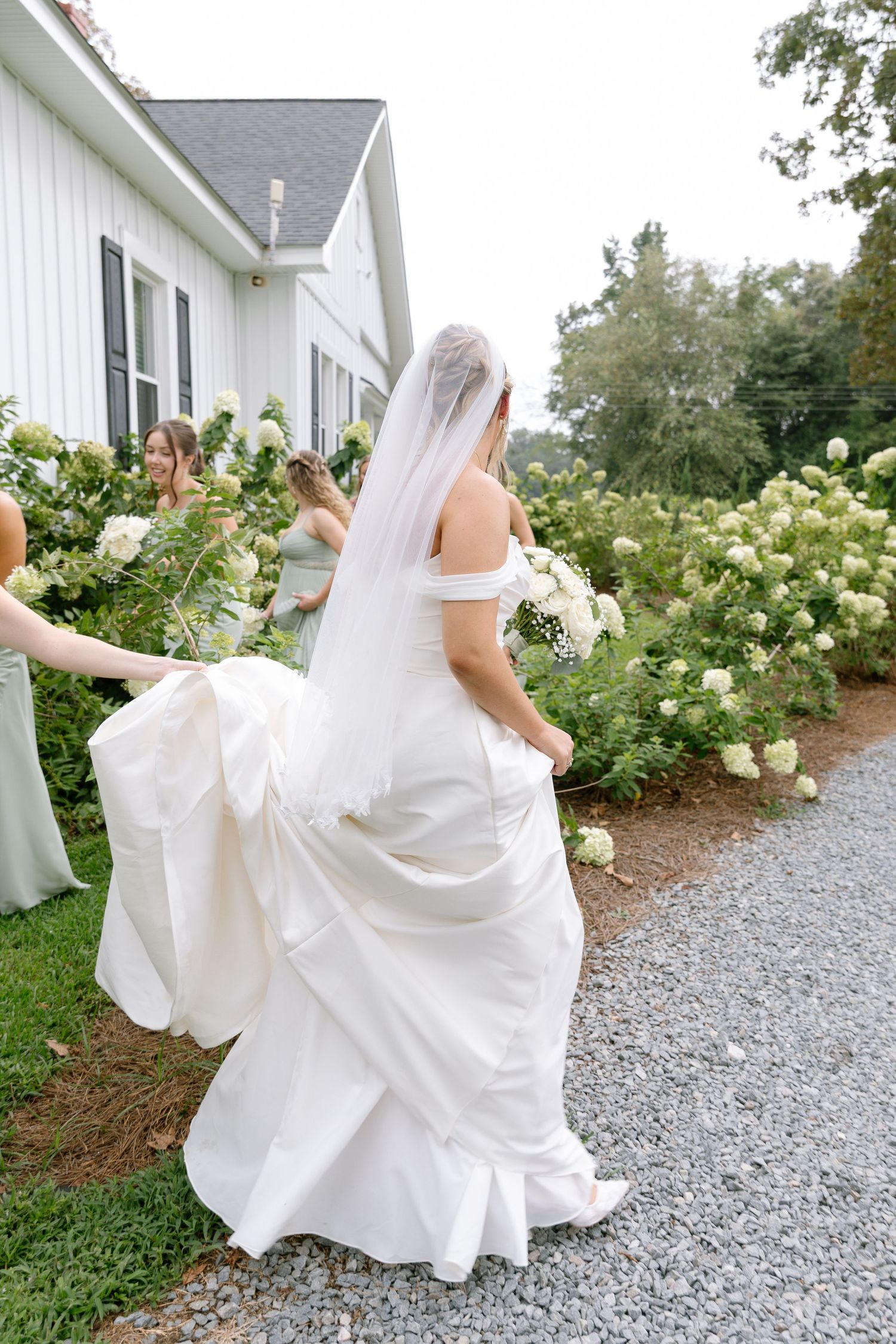 Bride in elegant white gown with veil walks path lined with white hydrangeas accompanied by bridesmaids.