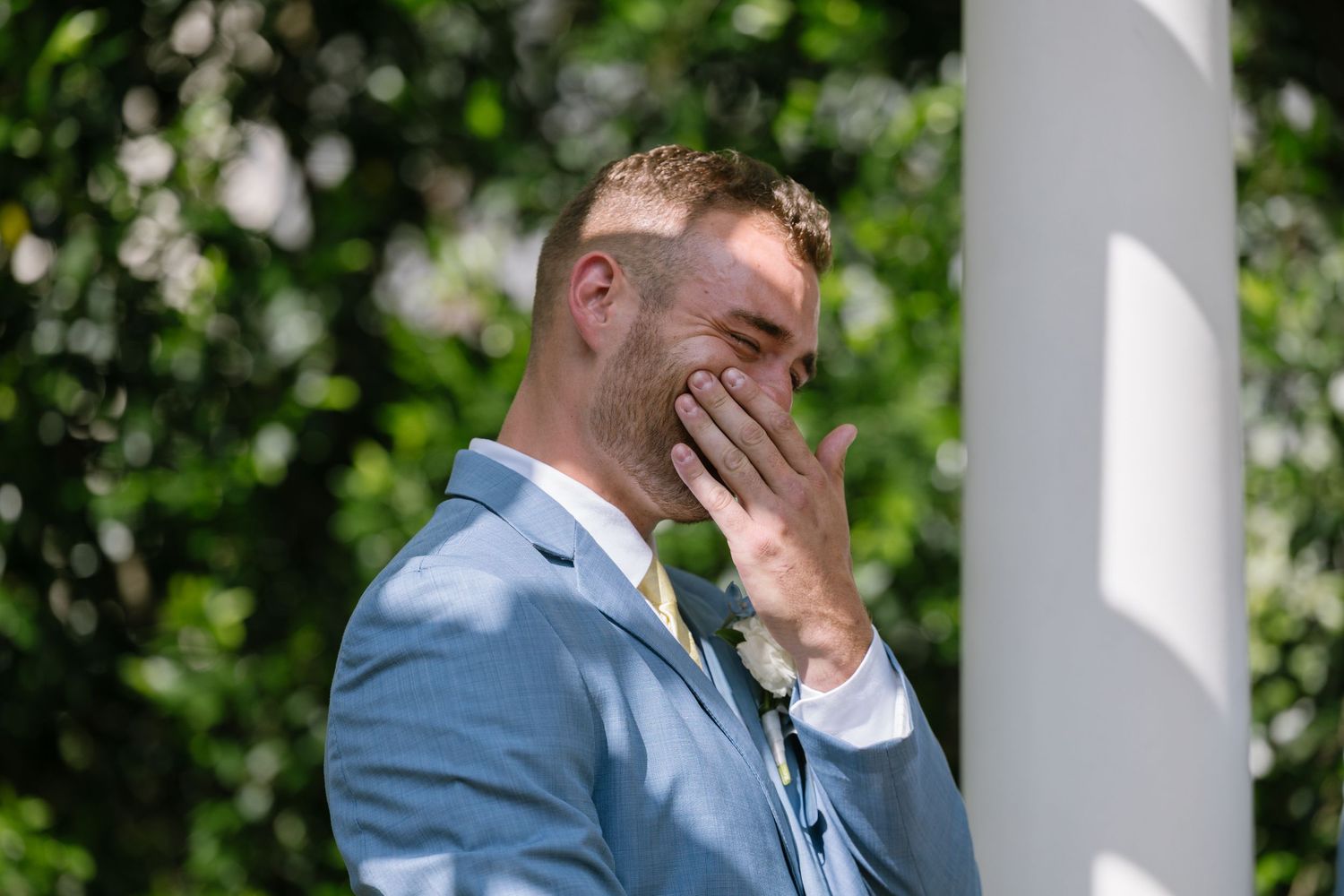 Emotional groom in blue suit covering his mouth with hand during outdoor wedding ceremony.