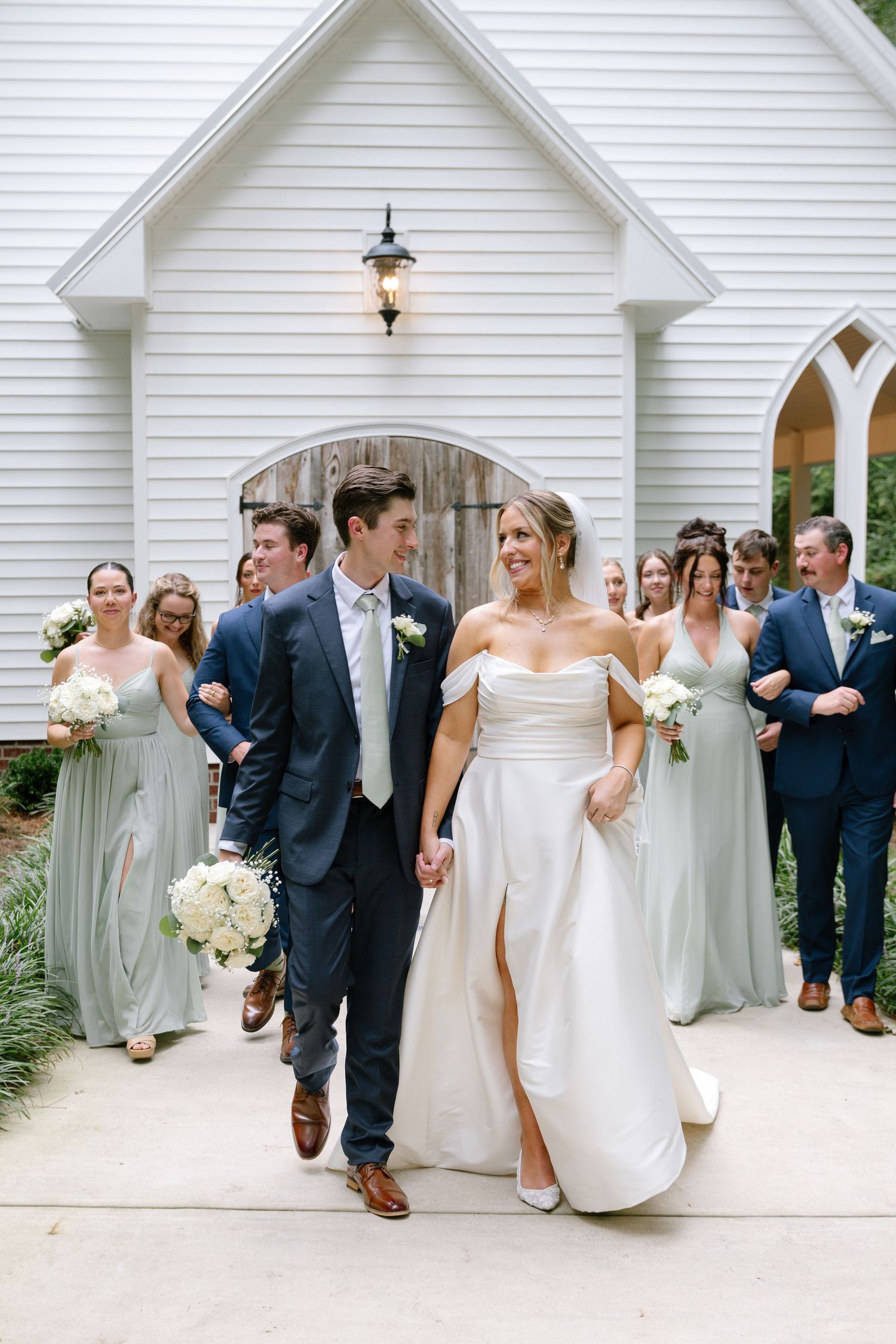 Joyful bride and groom leading wedding party outside a white chapel after the ceremony.