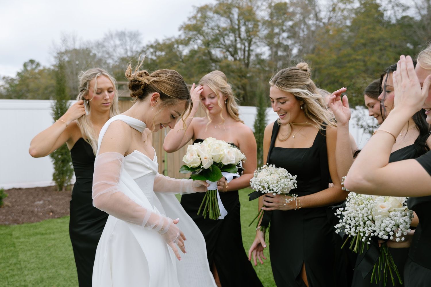 Bride in white gown laughs with bridesmaids in black dresses holding white bouquets outdoors.