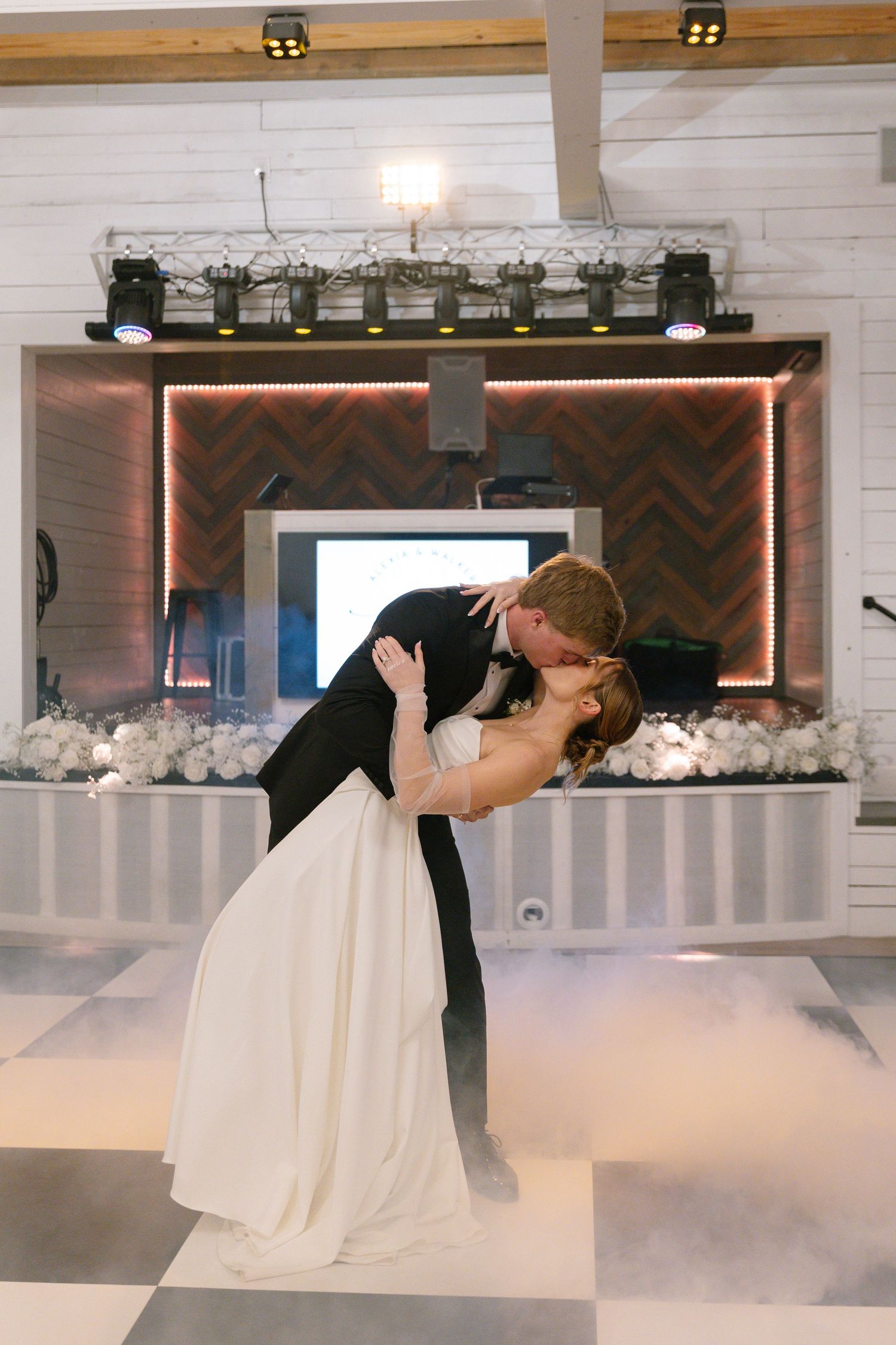 Bride and groom sharing romantic dip kiss on foggy black and white checkered dance floor.