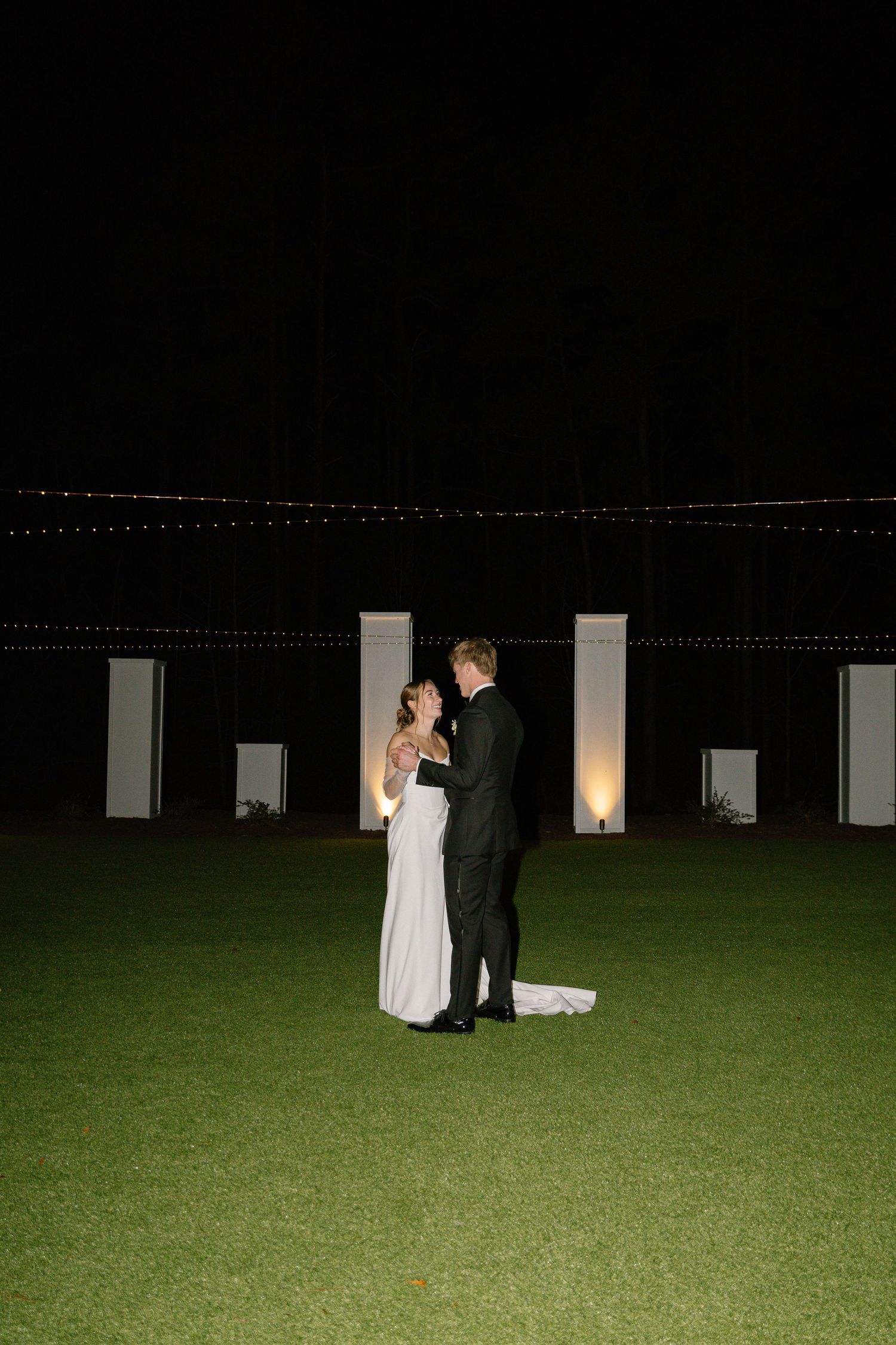 Bride and groom sharing a romantic embrace on a green lawn under string lights at night.