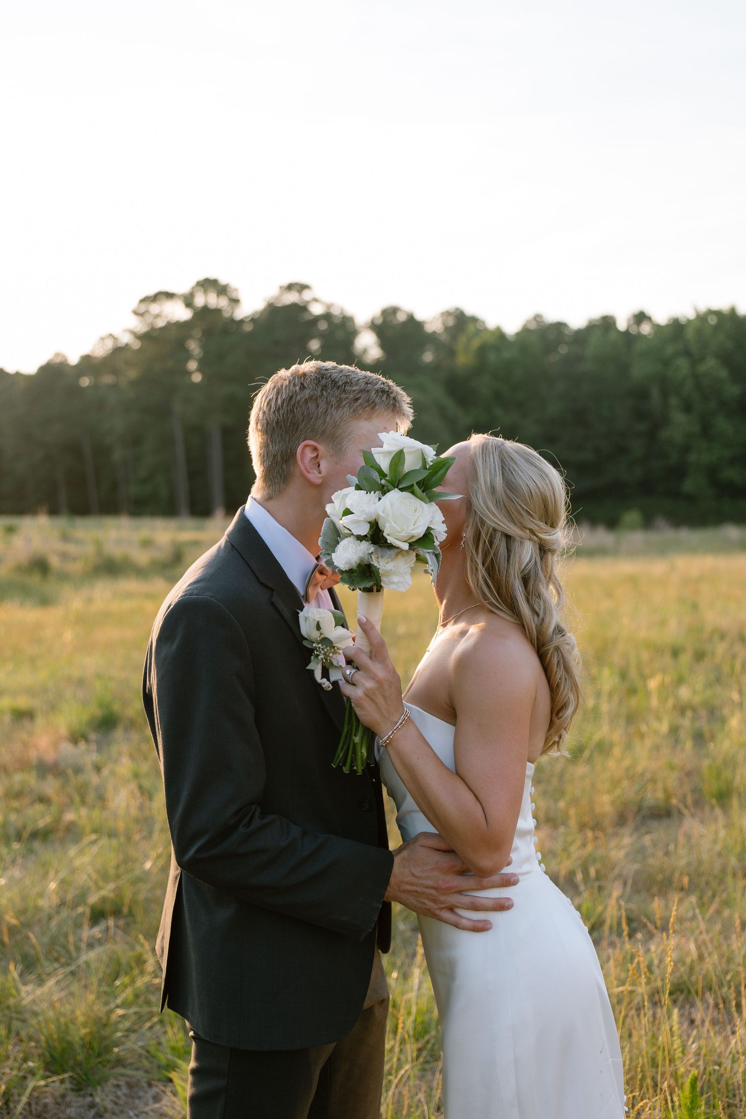 Bride and groom kissing behind white floral bouquet in golden hour sunlit field with trees in background.