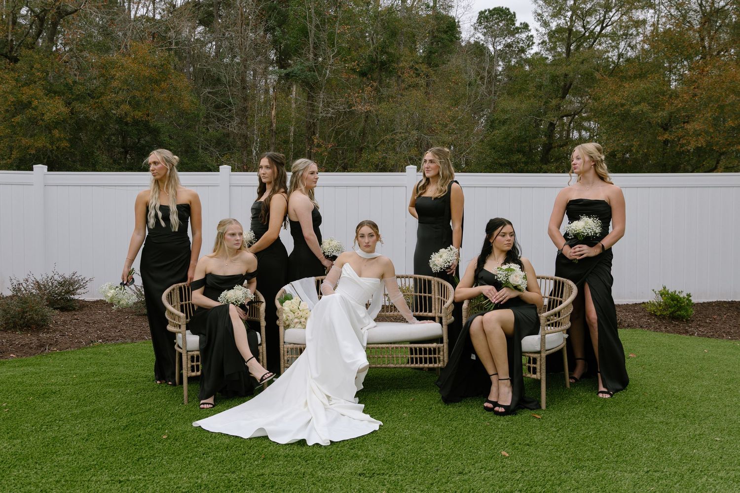 Bridesmaids in black gowns pose with bride in white dress on garden lawn with gold chairs.