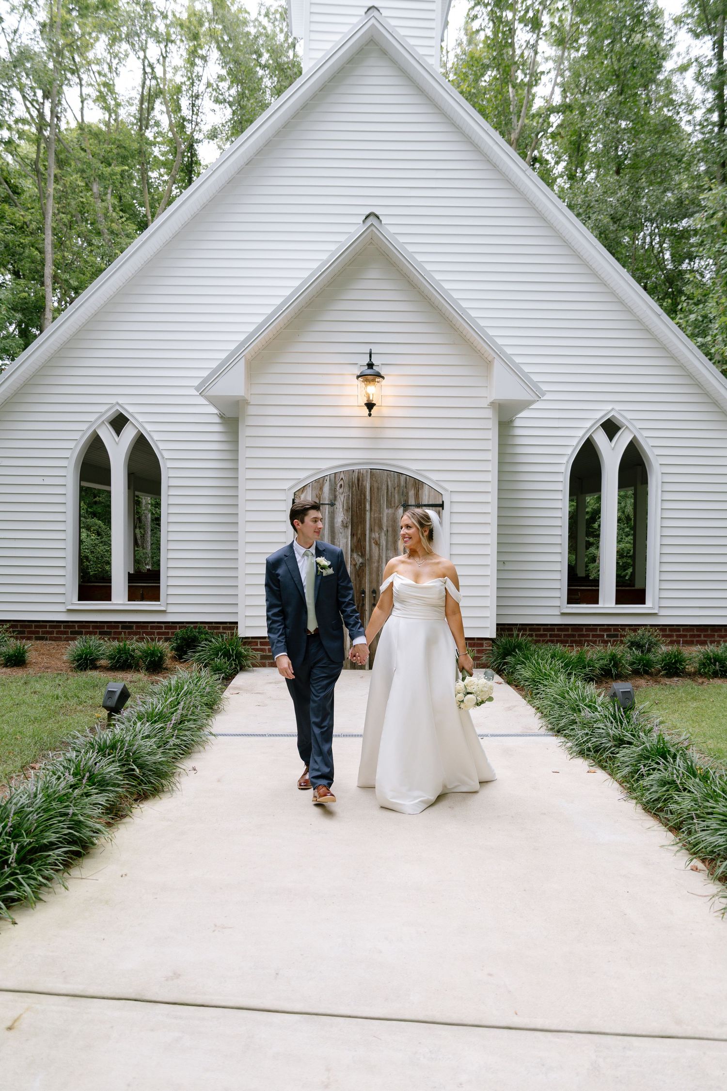 Bride in off-shoulder gown and groom in navy suit walking in front of a white chapel wedding venue.