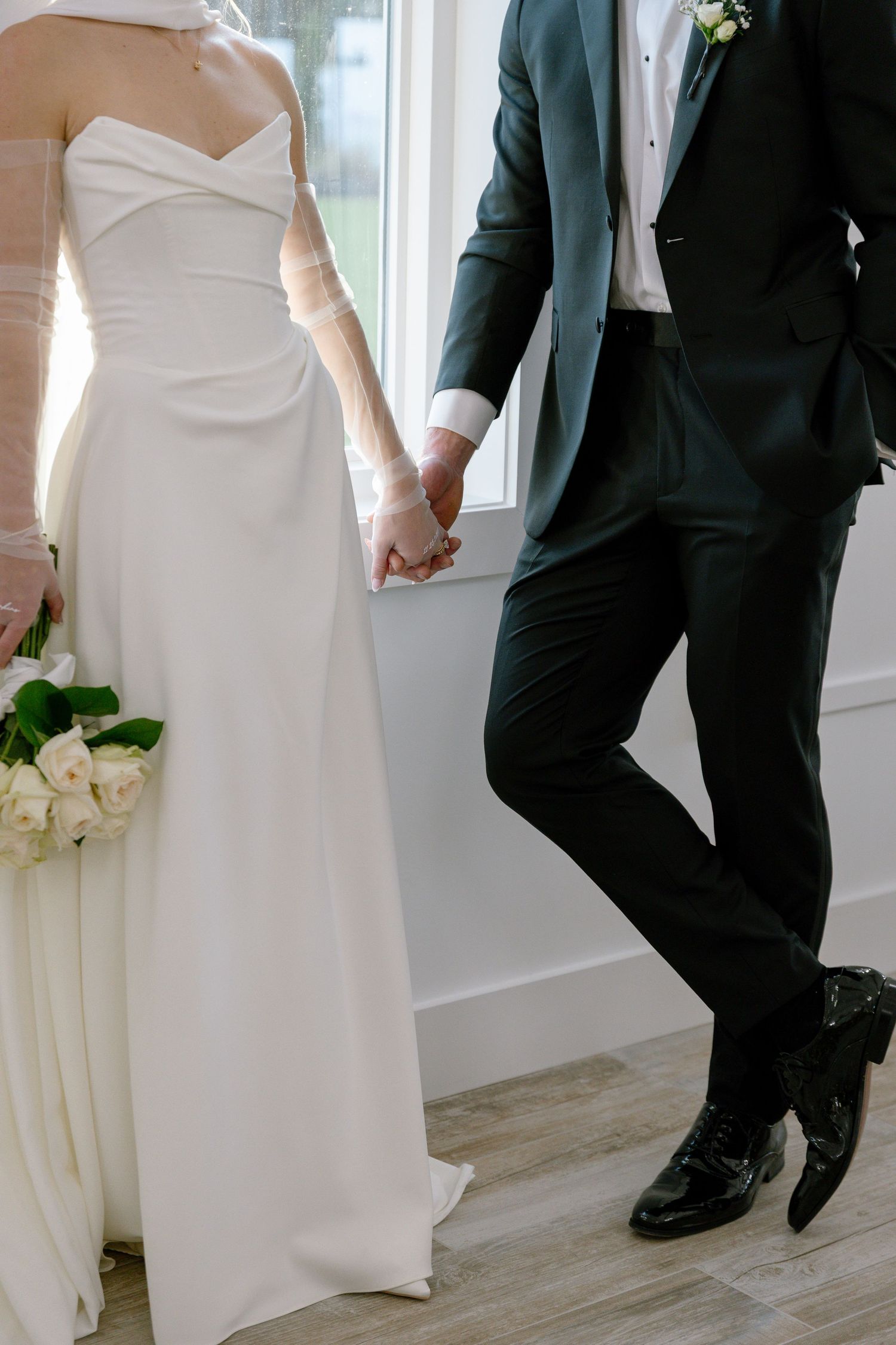 Bride in elegant strapless white gown holds hands with groom in dark suit while holding white rose bouquet.