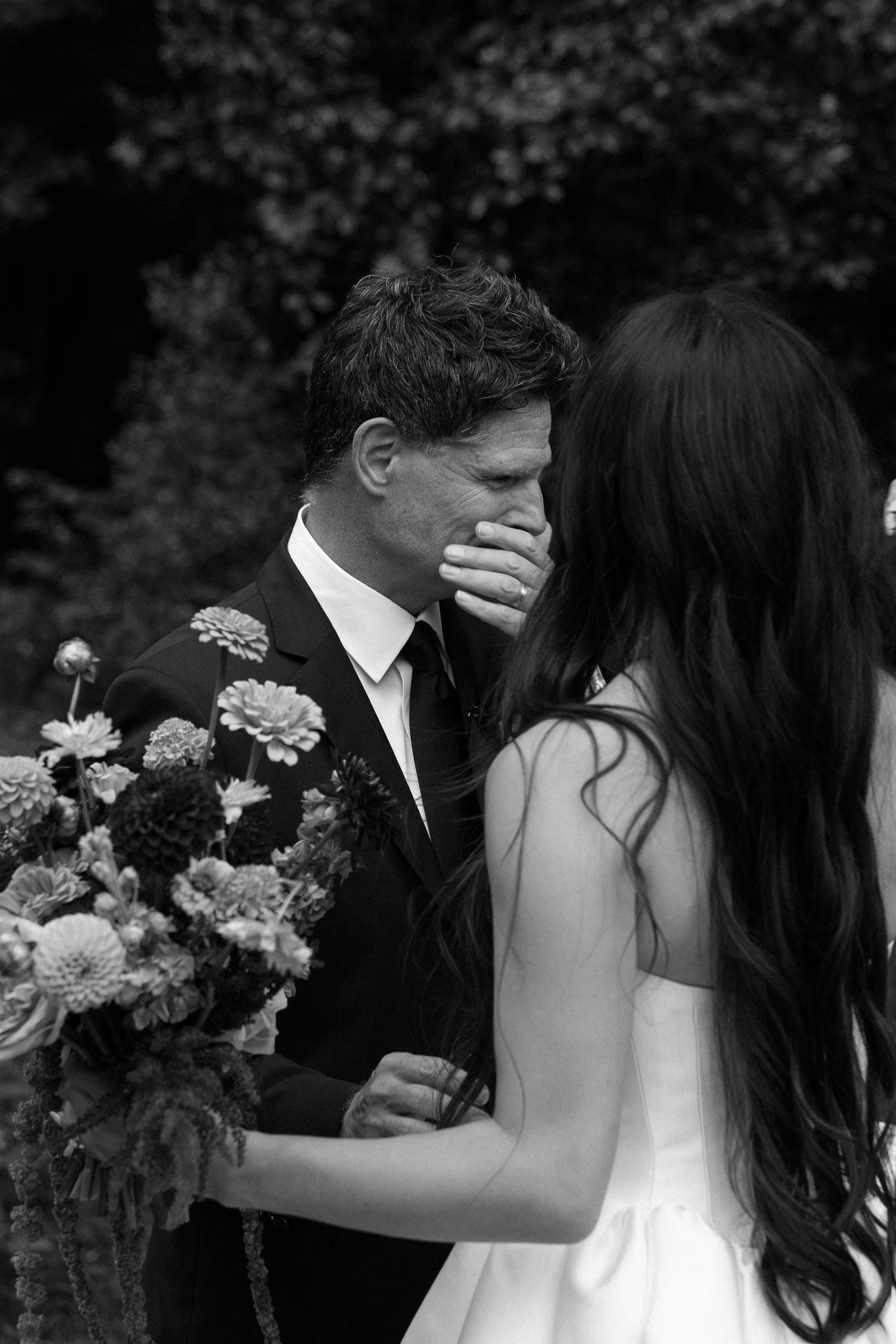 Black and white photo of emotional groom covering his mouth as bride faces him holding a large floral bouquet at wedding.