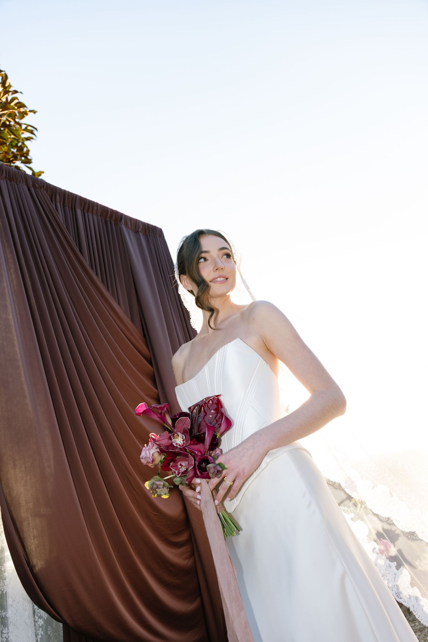 Bride in white gown holds burgundy floral bouquet beside draped brown fabric with scenic coastal backdrop.