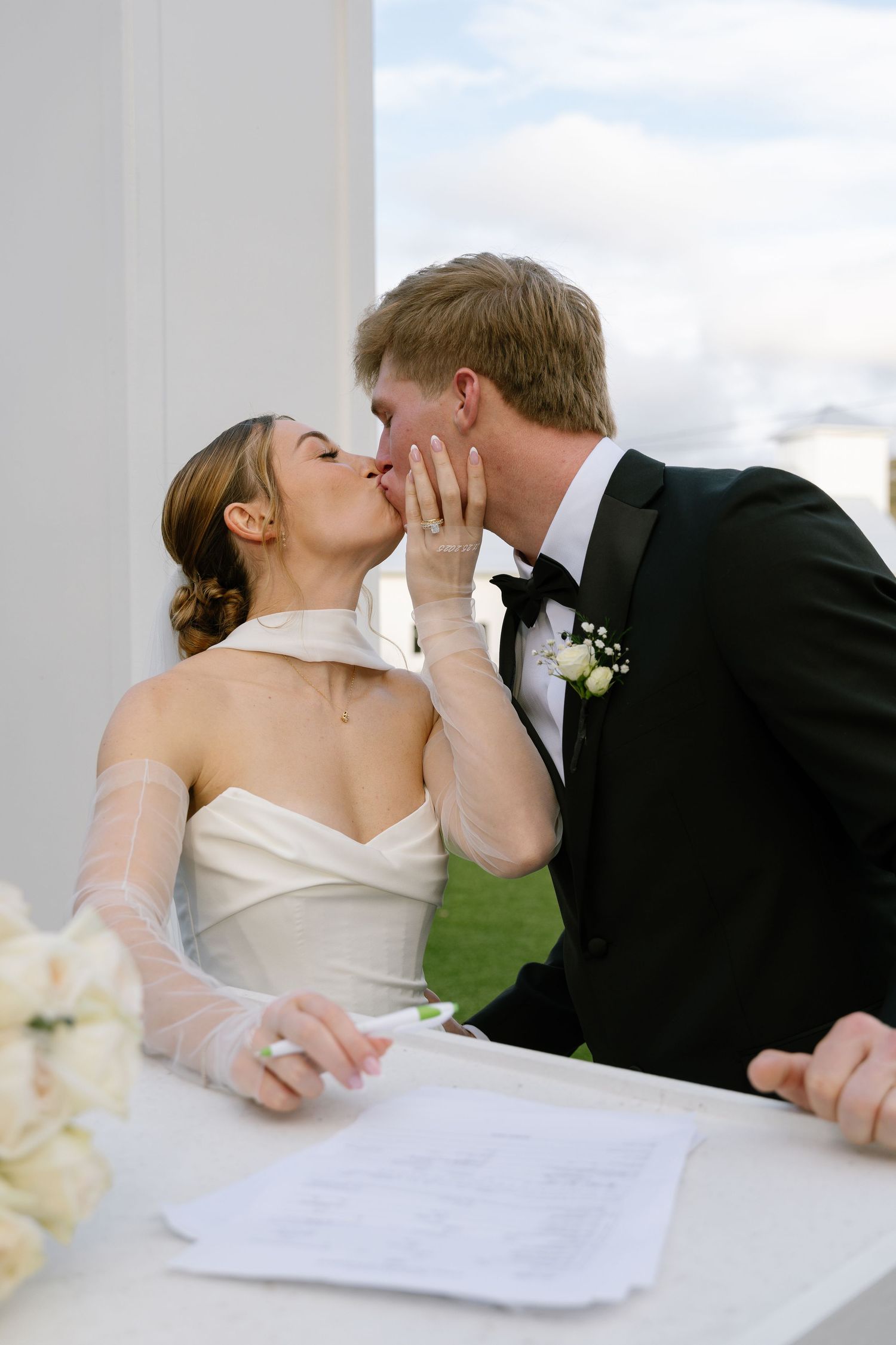 Bride and groom share a kiss while signing wedding registry at outdoor ceremony.