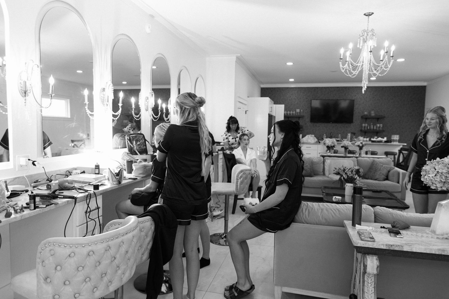 Black and white photo of women getting hair and makeup done in a bright, elegant salon with chandeliers.