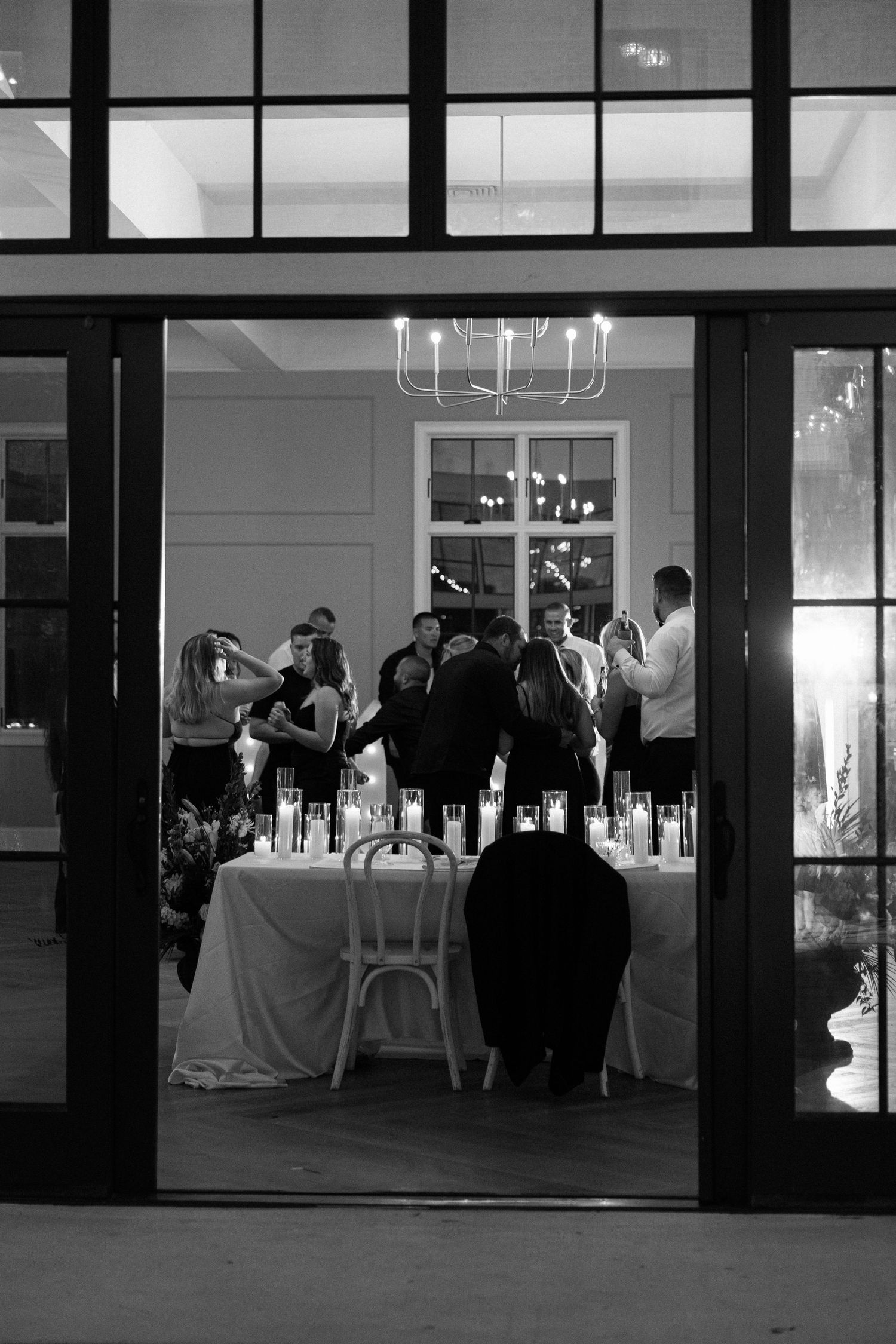 Black and white wedding reception photo showing guests gathered around candlelit tables inside an elegant venue with glass doors.