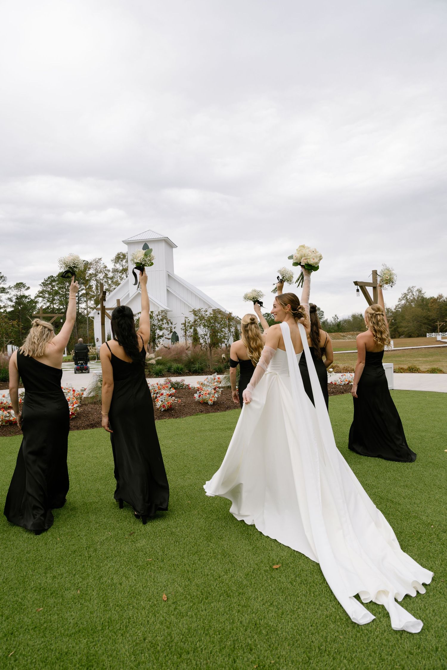 Bride and bridesmaids in black gowns raise white bouquets toward white chapel on cloudy day.
