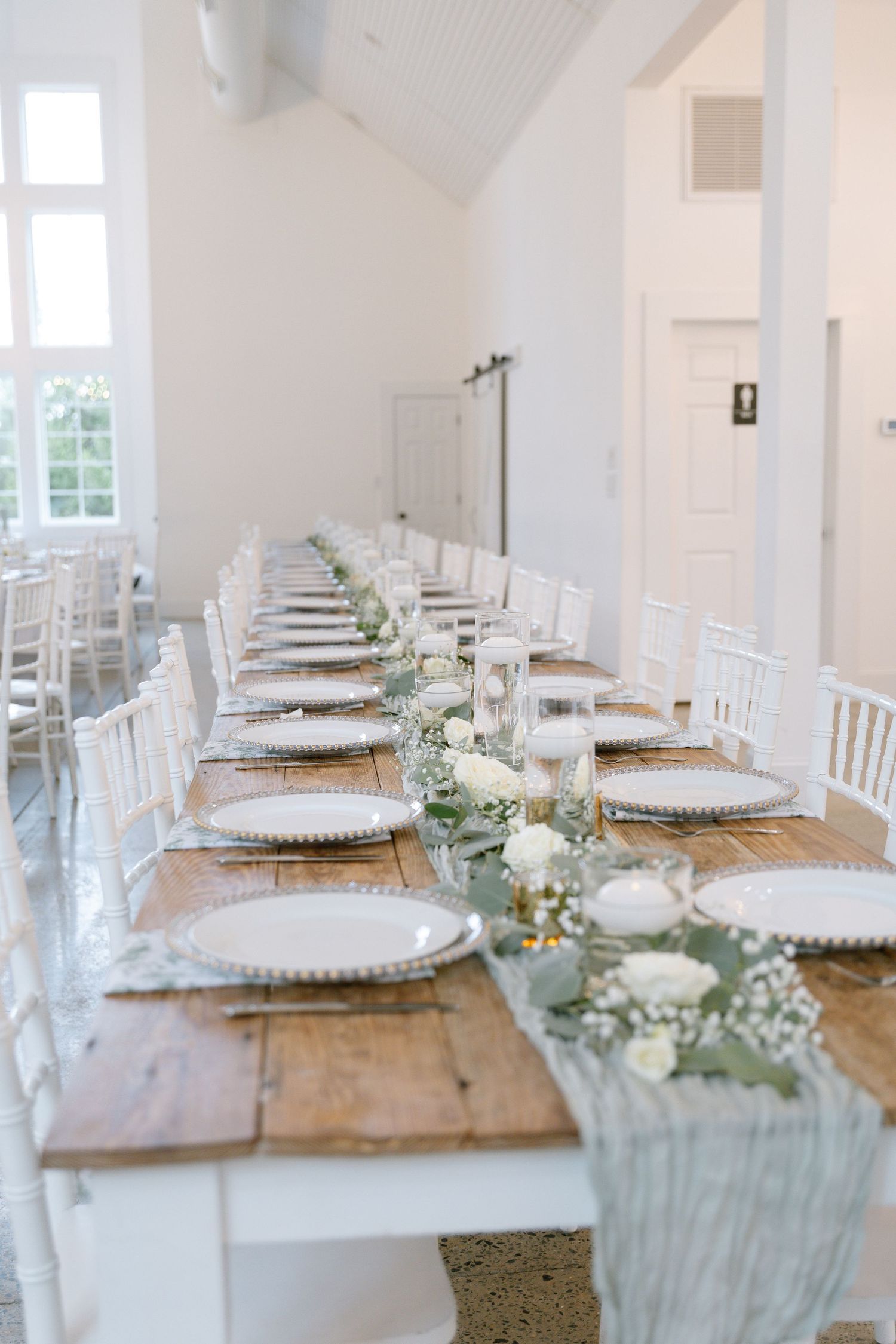 Long rustic farm table with white chiavari chairs, silver charger plates, and greenery floral runner for a wedding reception.