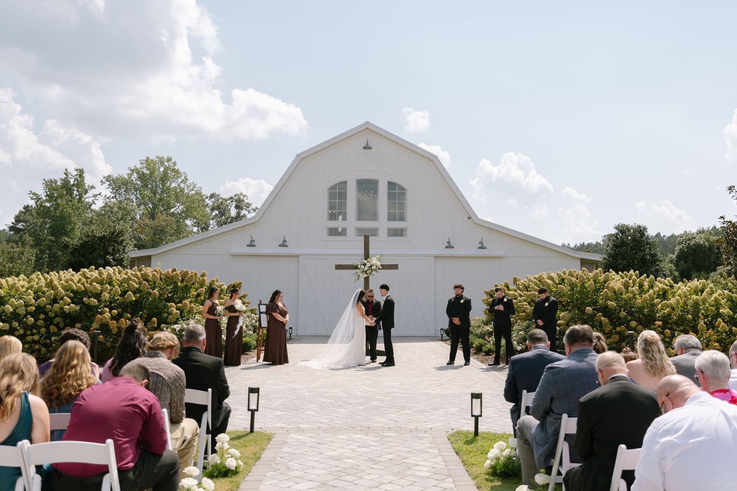 Outdoor wedding ceremony in front of a white barn venue with guests seated on white chairs under a sunny sky.