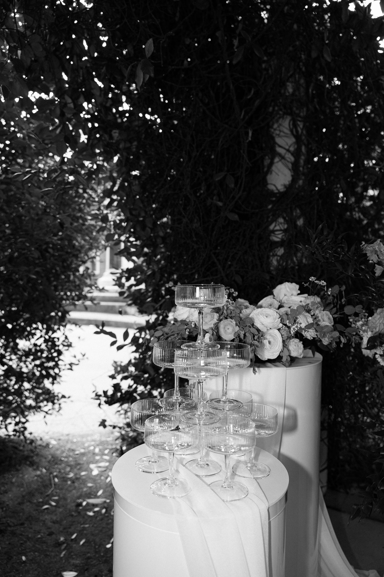 Black and white photo of champagne tower with floral arrangements on draped table, surrounded by lush greenery.