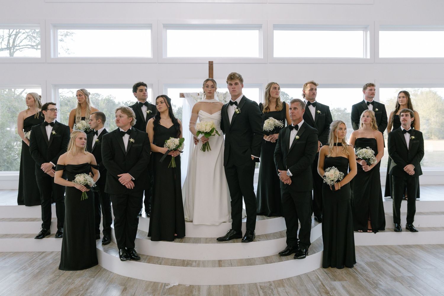 Wedding party in black attire with bride and groom centered on steps in elegant white venue.