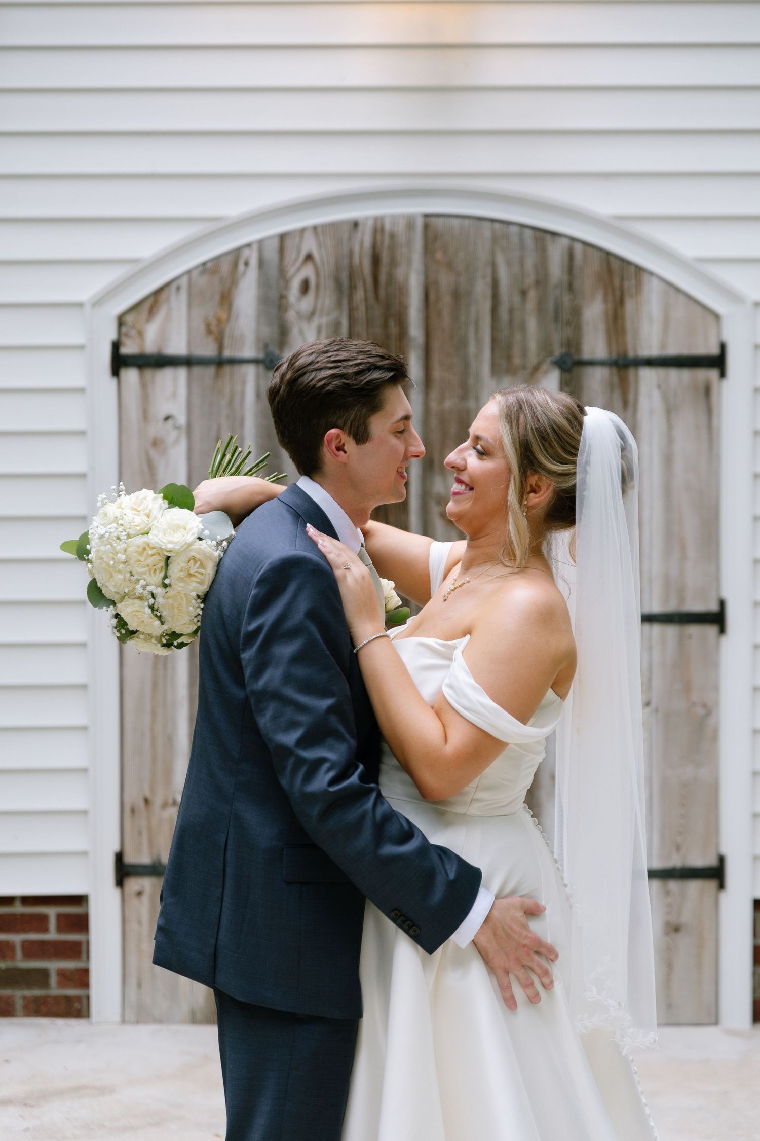 Bride and groom share a romantic moment embracing outside rustic wooden doors, bride holding white hydrangea bouquet.
