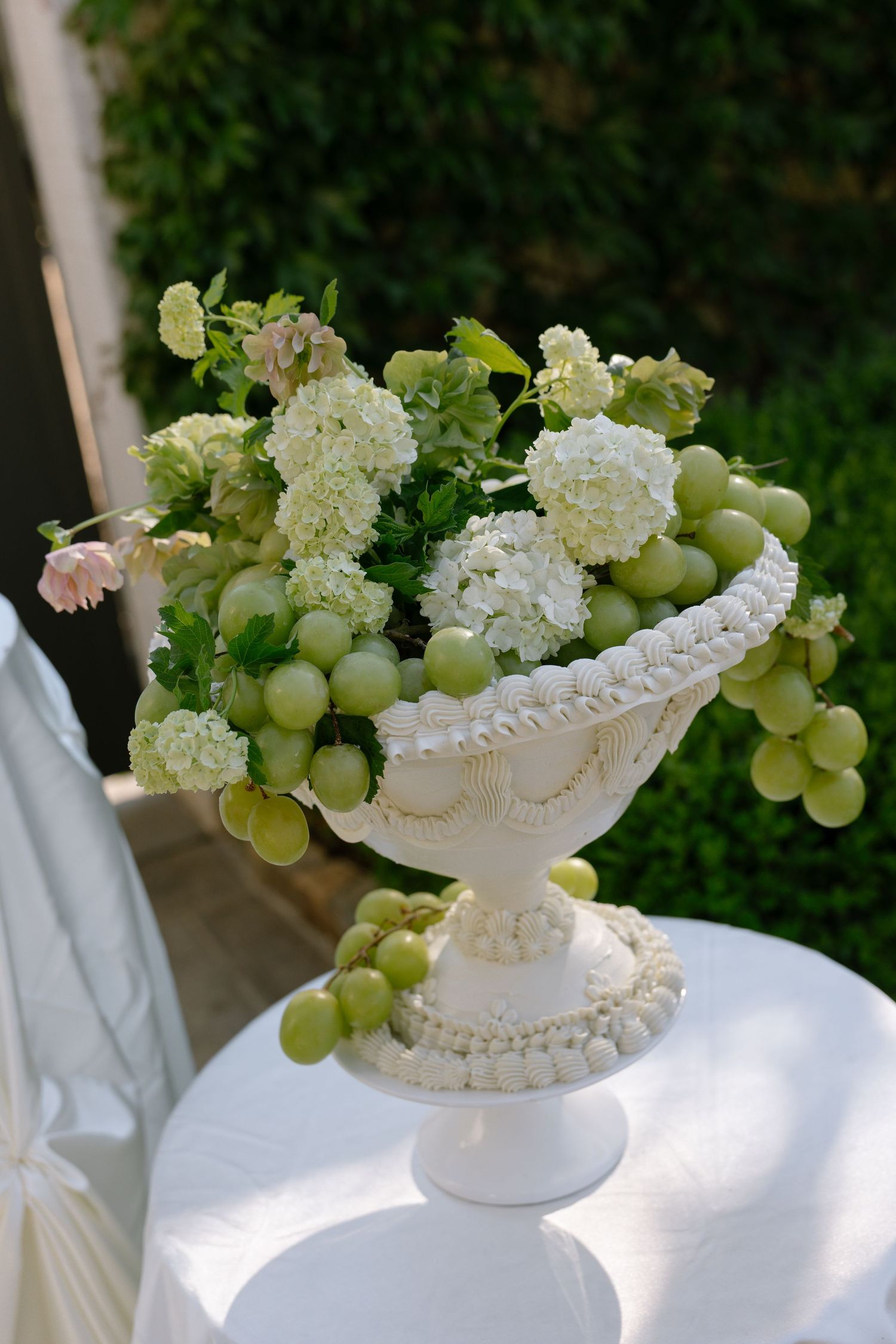Elegant white ceramic pedestal vase filled with green grapes and white hydrangea flowers on a draped table outdoors.
