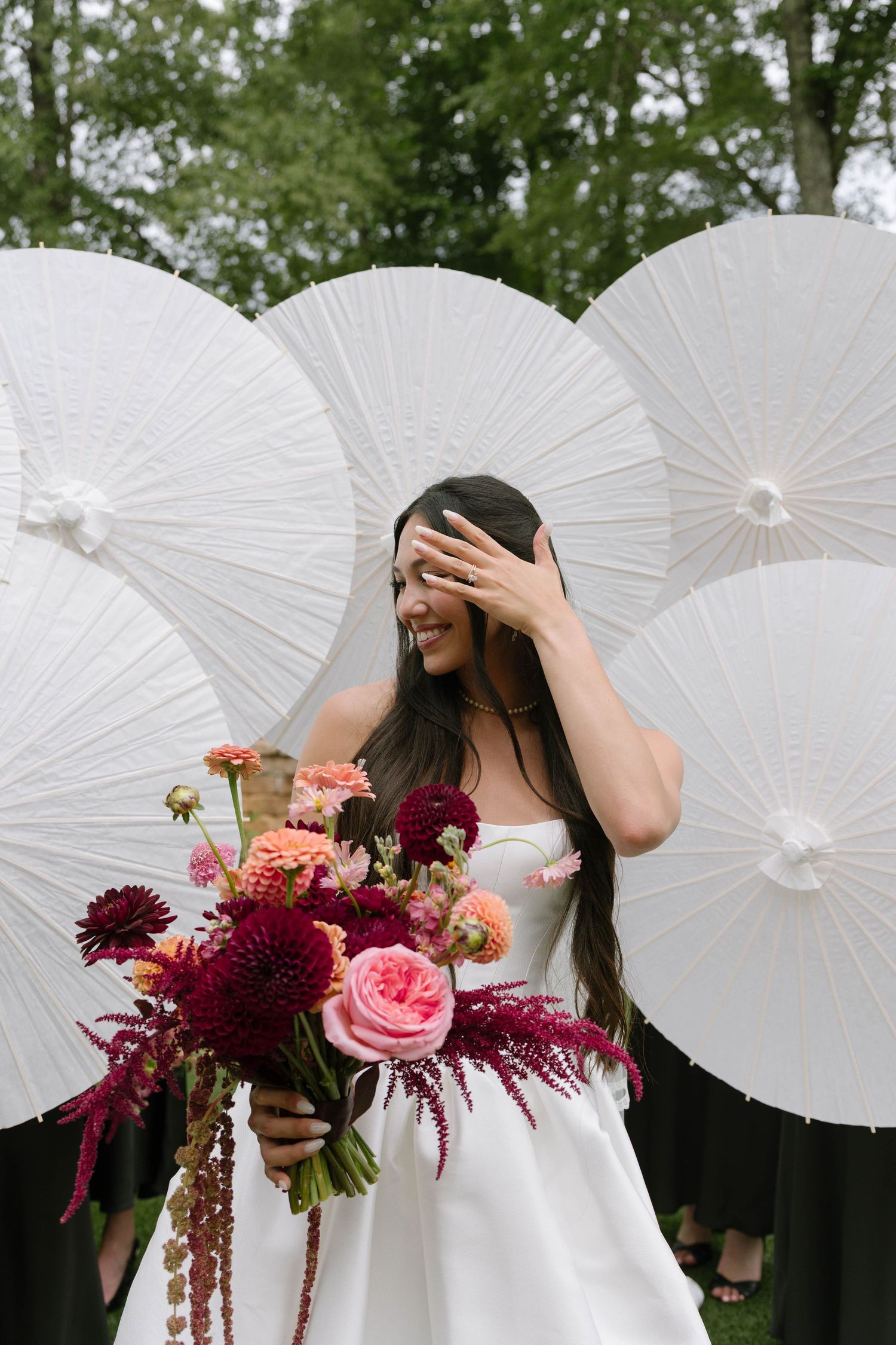 Smiling bride in white gown holds colorful bouquet of dahlias and roses, standing before white paper parasols outdoors.