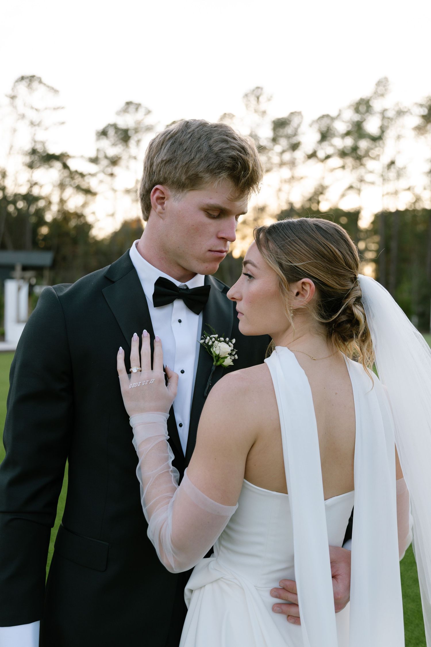 Bride and groom in black tuxedo and white wedding gown share a romantic moment outdoors at golden hour.