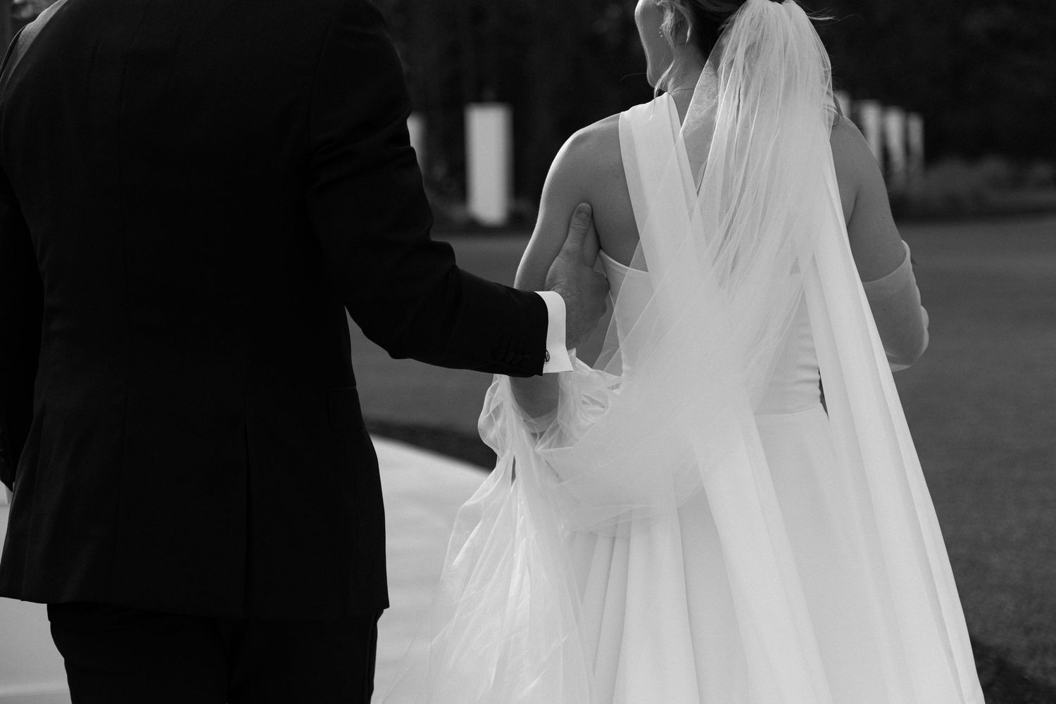Black and white photo of bride and groom walking together, showcasing flowing wedding veil.
