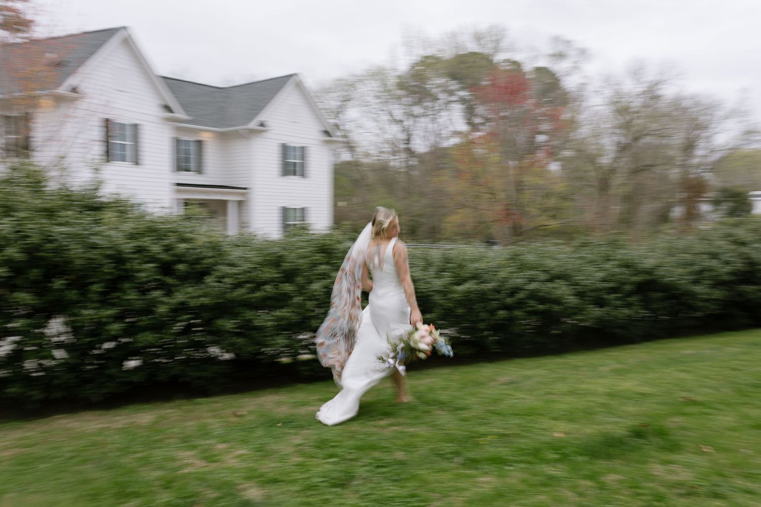 Bride in white gown and veil runs across green lawn near white farmhouse on cloudy autumn wedding day.