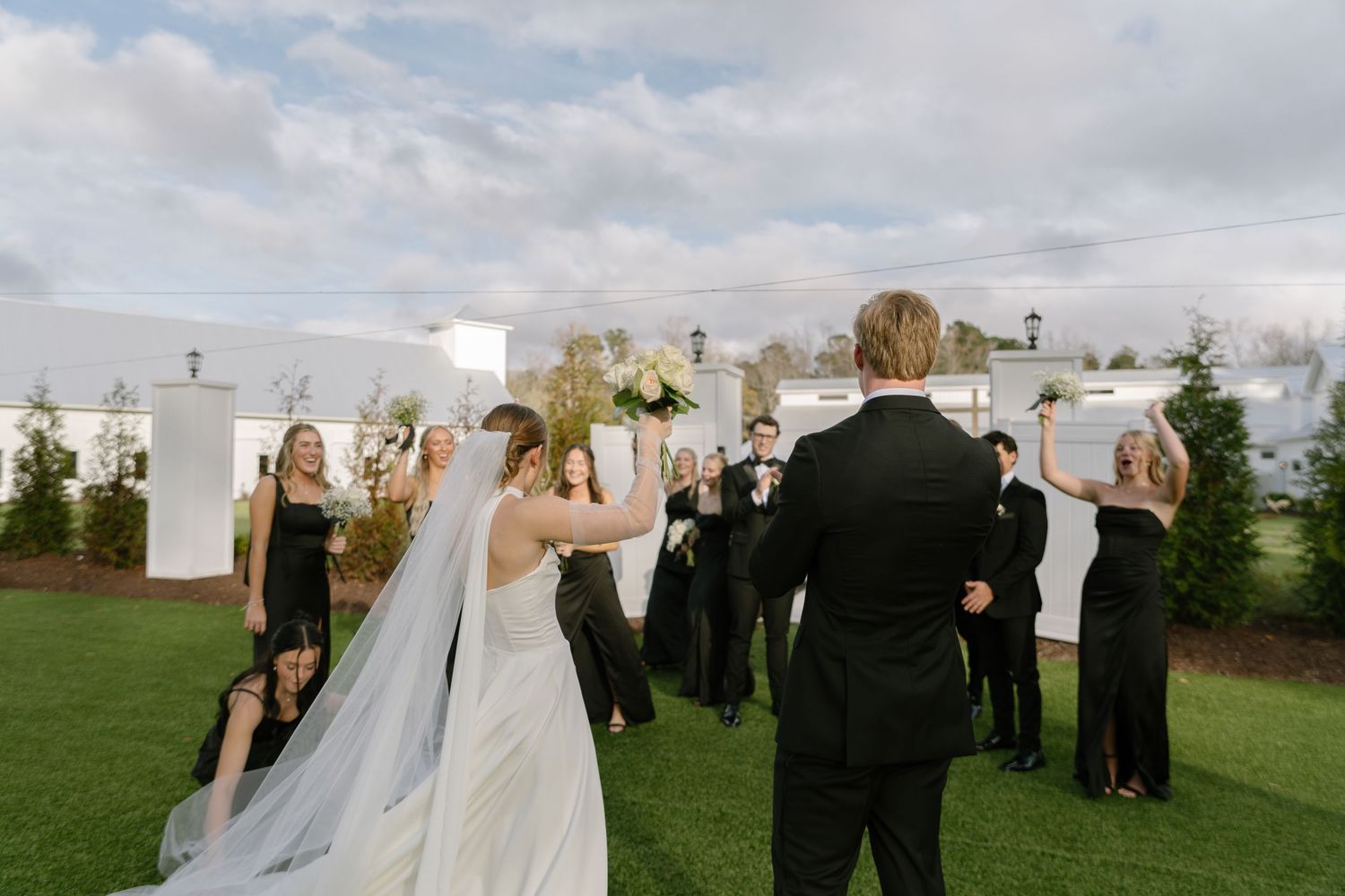 Bride and groom celebrate with wedding party on green lawn, bridal party dressed in black attire.