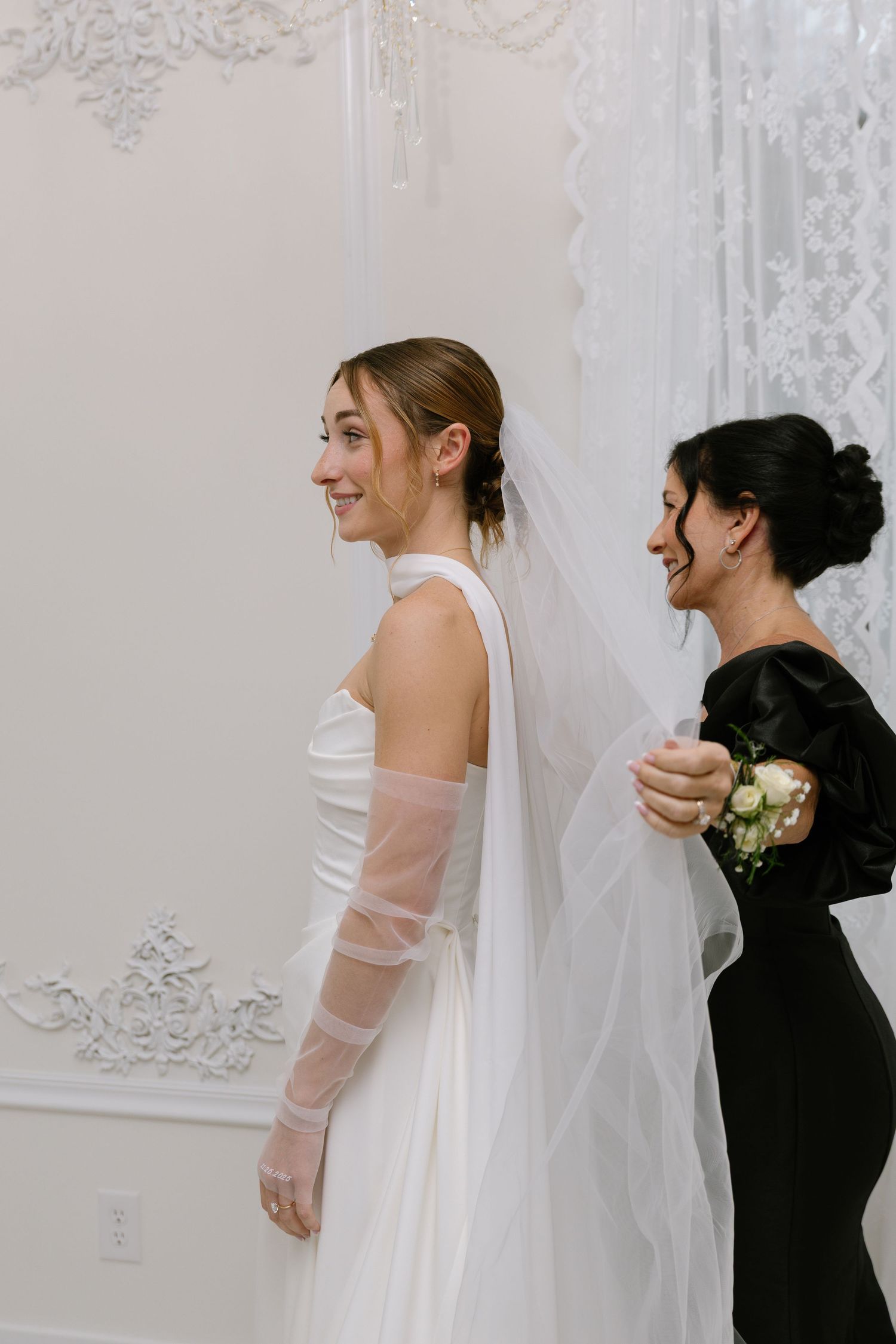 Bride in white halter gown with sheer sleeves smiles as bridesmaid in black dress adjusts her veil.