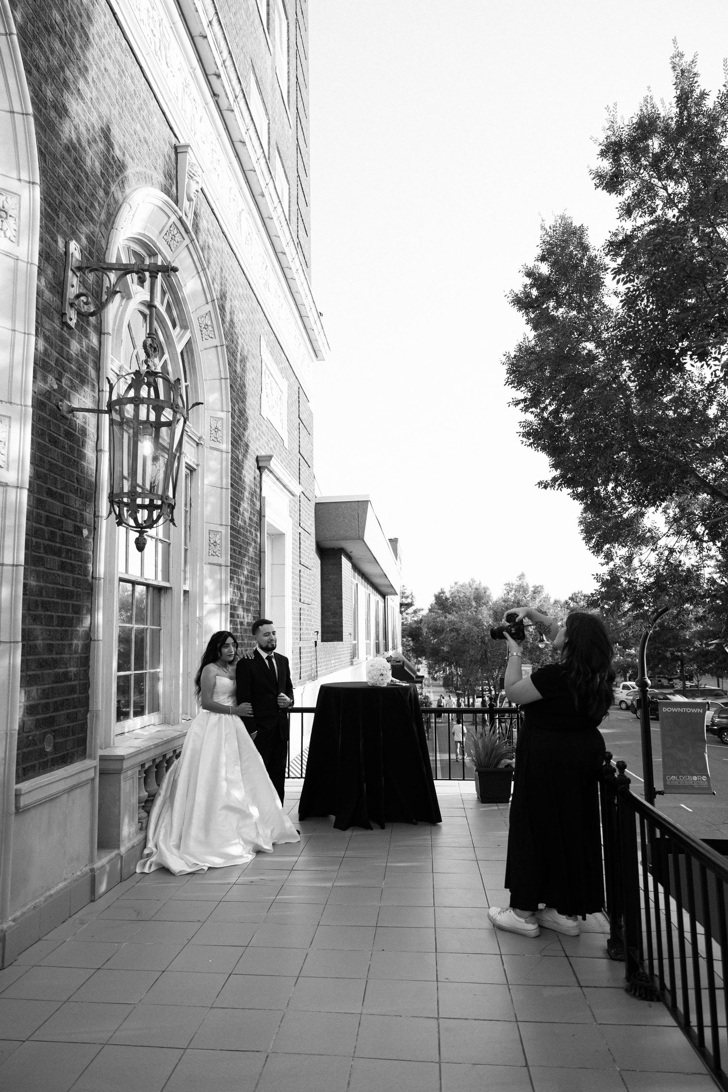 Black and white photo of a bride and groom posing outside a brick building while a photographer captures their wedding portrait.