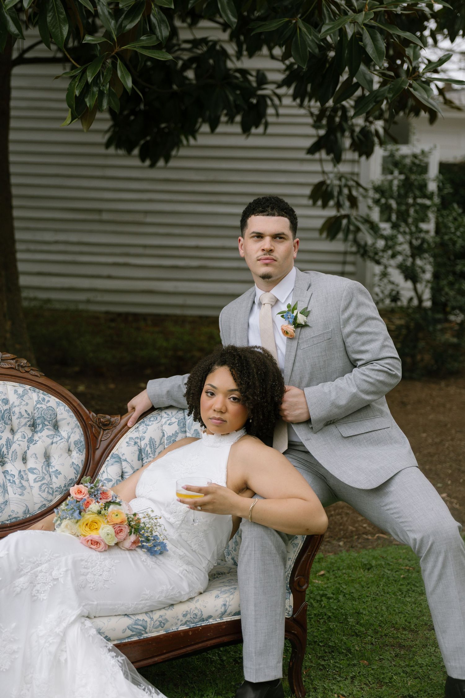 Bride in white gown sits on vintage floral sofa holding bouquet while groom in gray suit stands behind her outdoors.