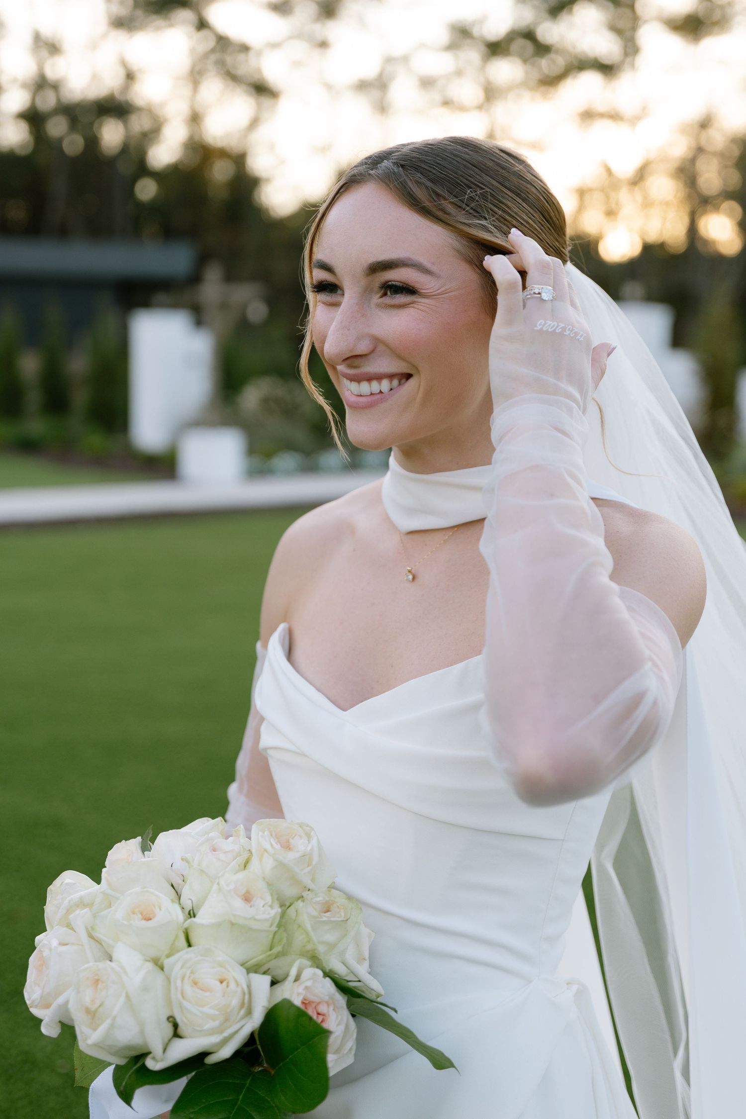 Smiling bride in white strapless gown with sheer gloves holds white rose bouquet at sunset.