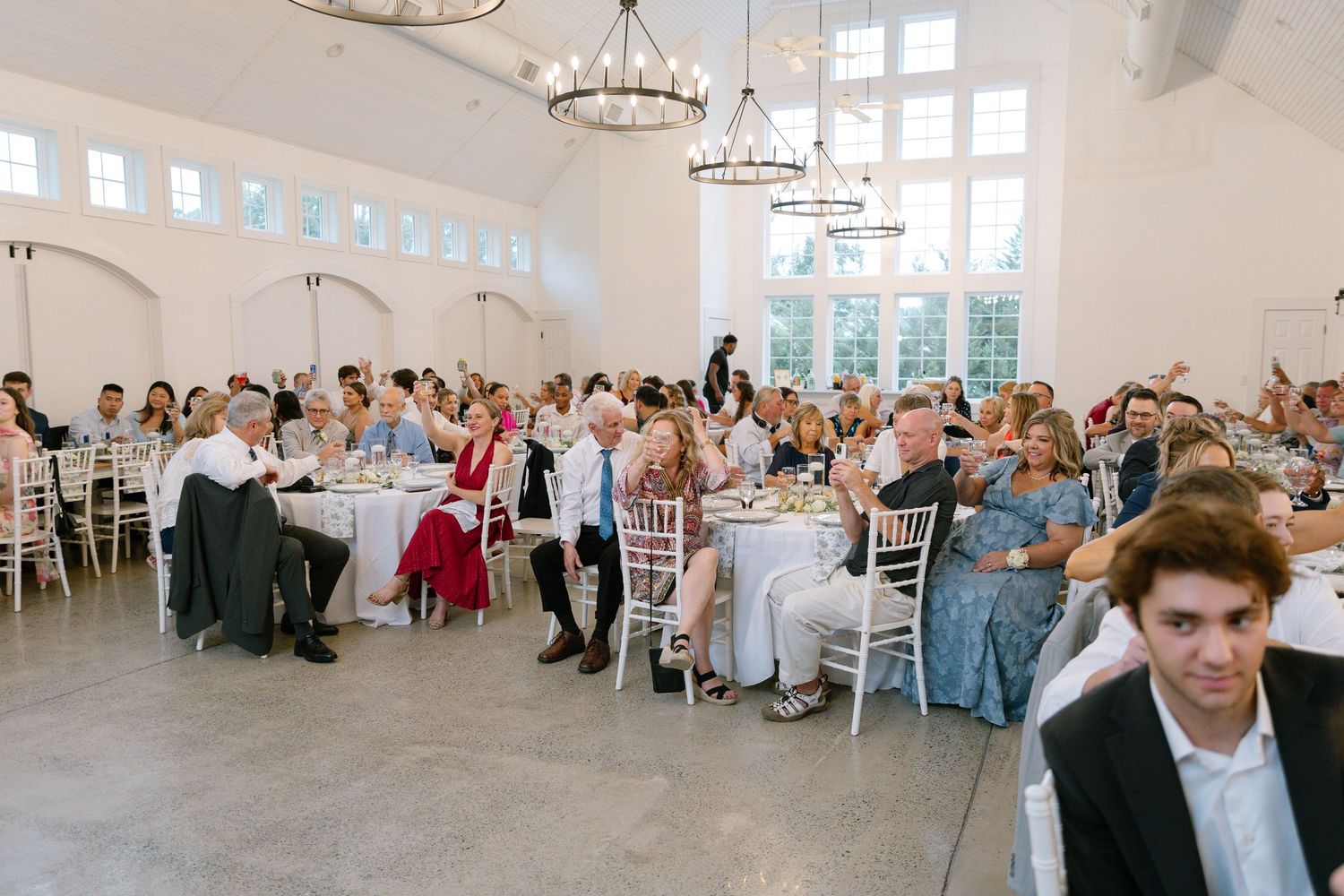 Wedding reception guests seated at round tables in a bright white hall with chandelier lighting.