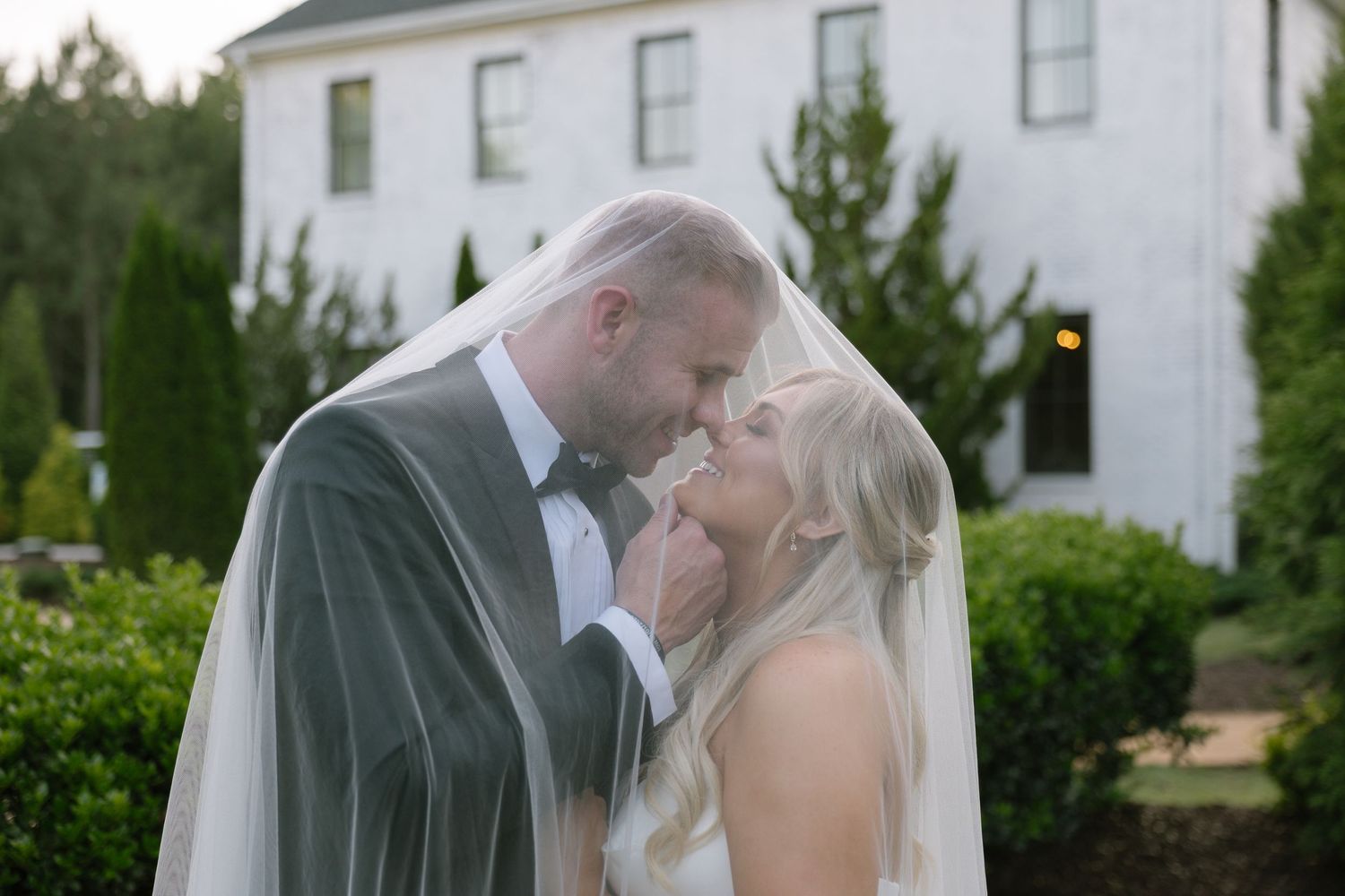 Bride and groom share romantic kiss under veil outdoors at elegant white wedding venue surrounded by lush greenery.