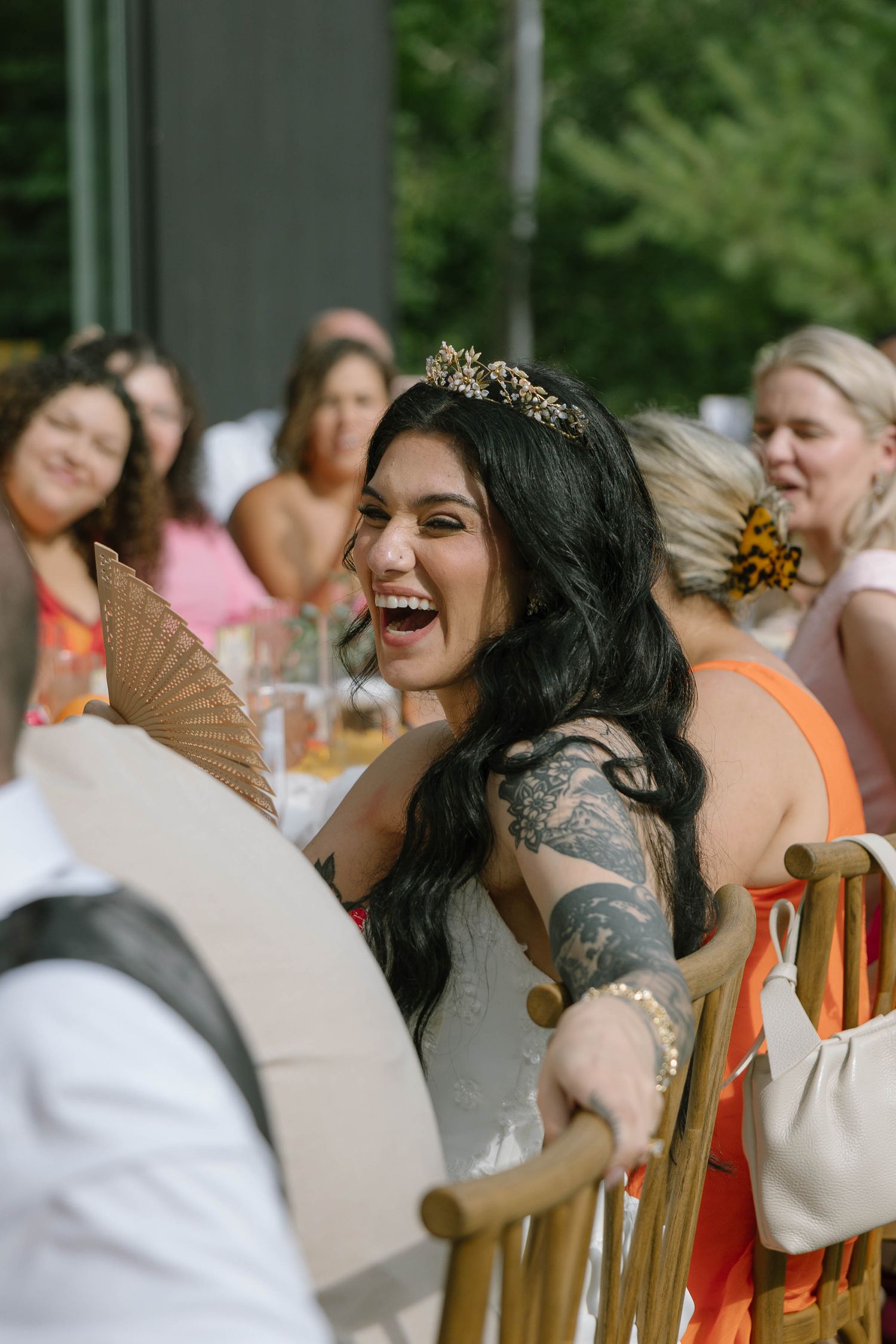 Laughing bride wearing a gold tiara and showing tattoos holds a fan at an outdoor wedding reception.