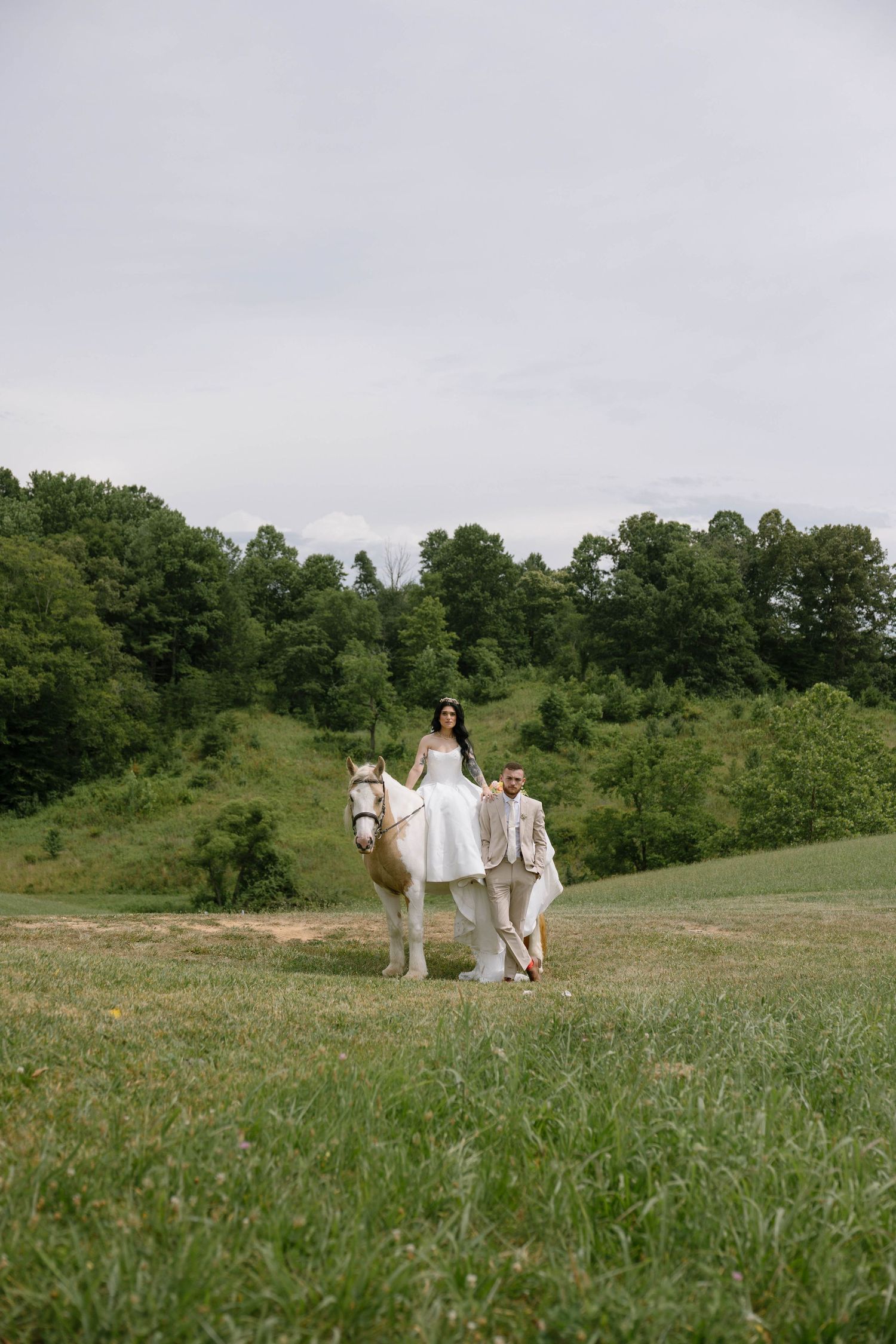 Bride in white dress riding a white horse led by groom in tan suit across a lush green field on their wedding day.