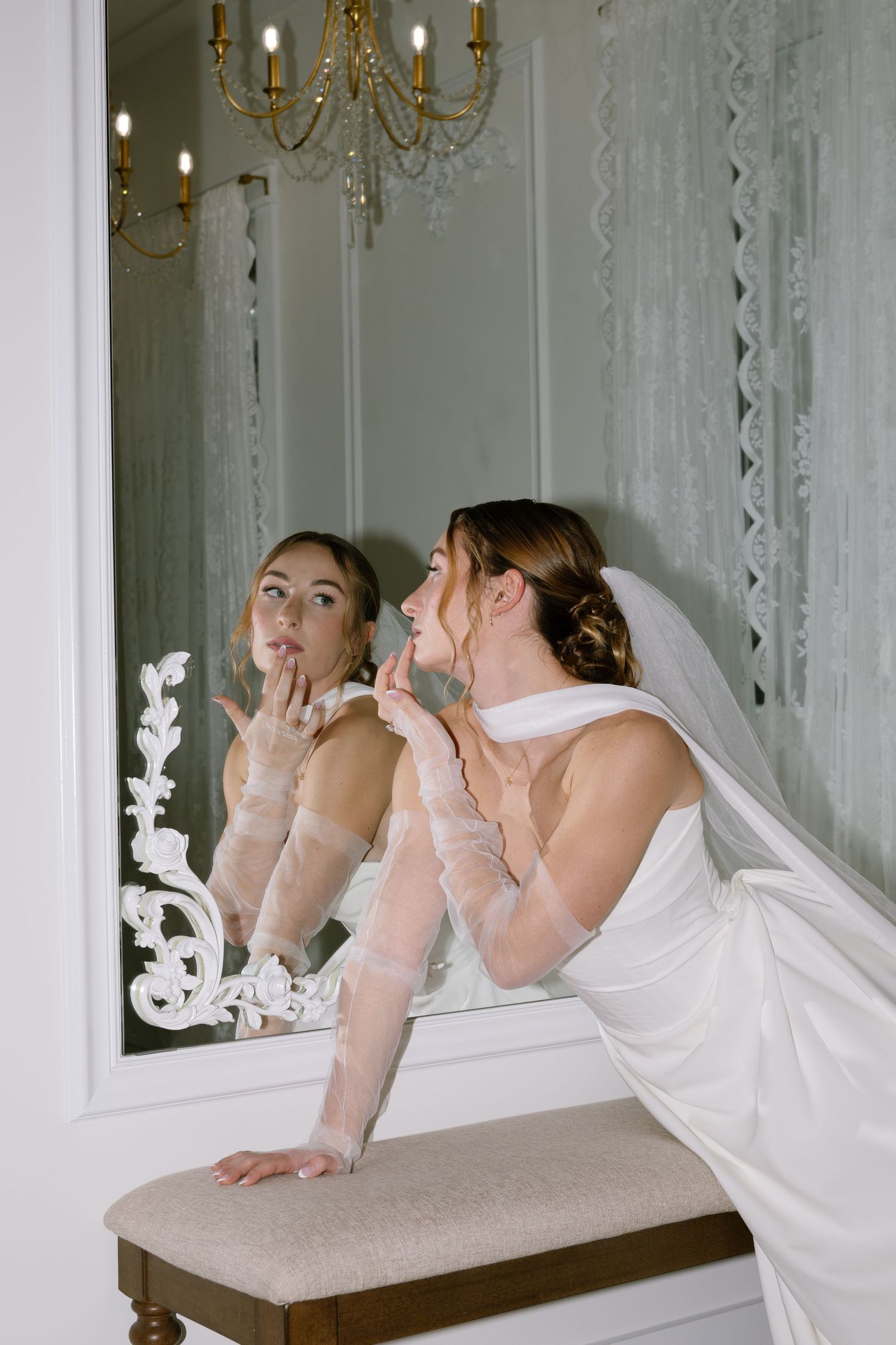 Bride in white gown and gloves touching up makeup while gazing into an ornate mirror with chandelier.