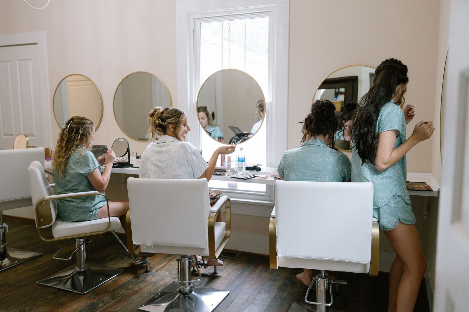 Bridesmaids getting hair and makeup done in a bright modern salon with round mirrors before a wedding.