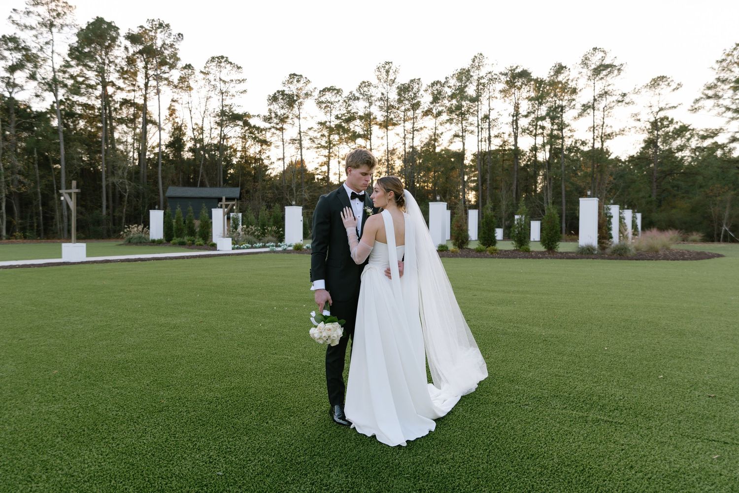 Bride and groom share a romantic moment on a lush green lawn surrounded by tall pine trees at sunset.