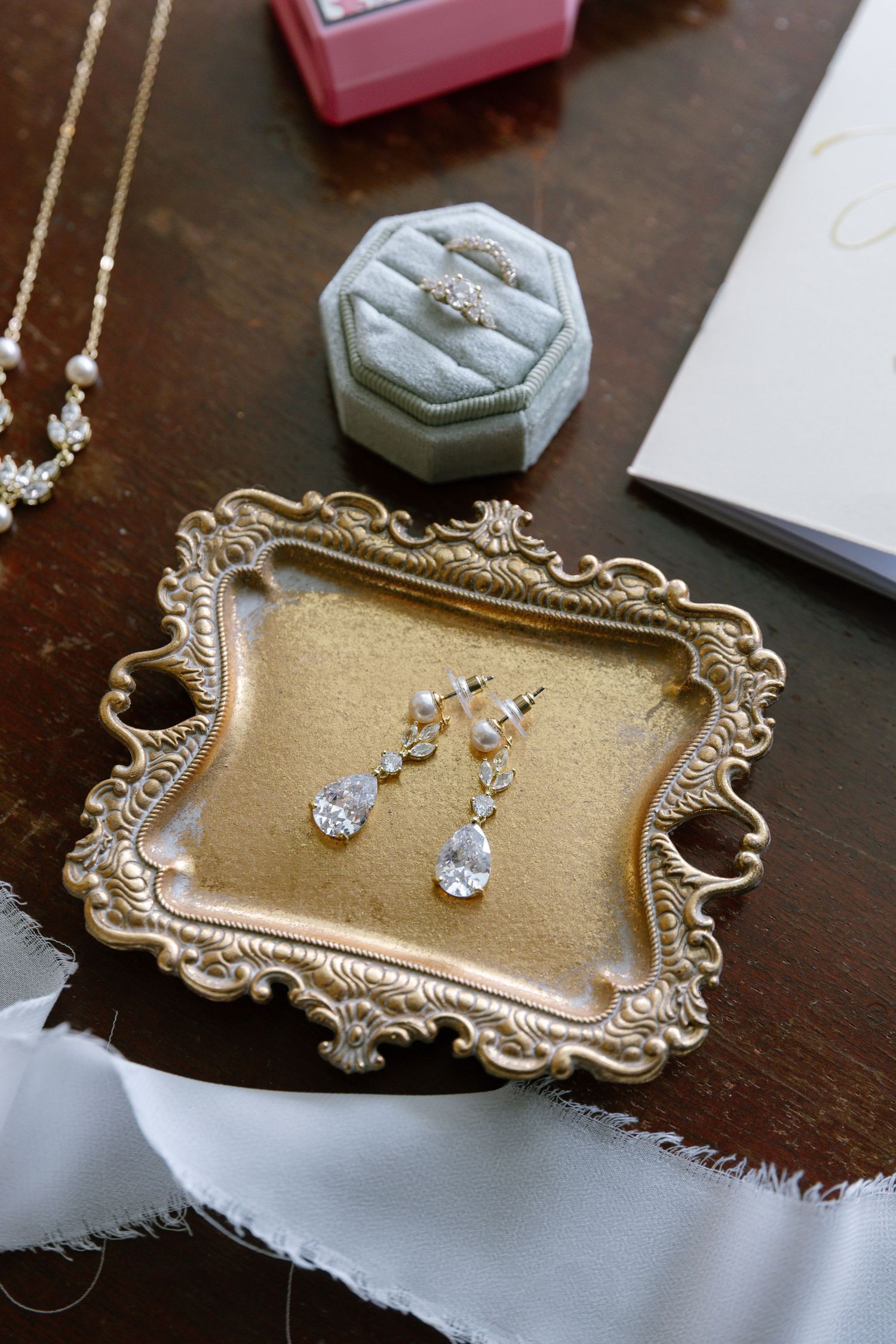 Elegant bridal jewelry flat lay featuring crystal drop earrings on a gold ornate tray with a ring box and pearl necklace.