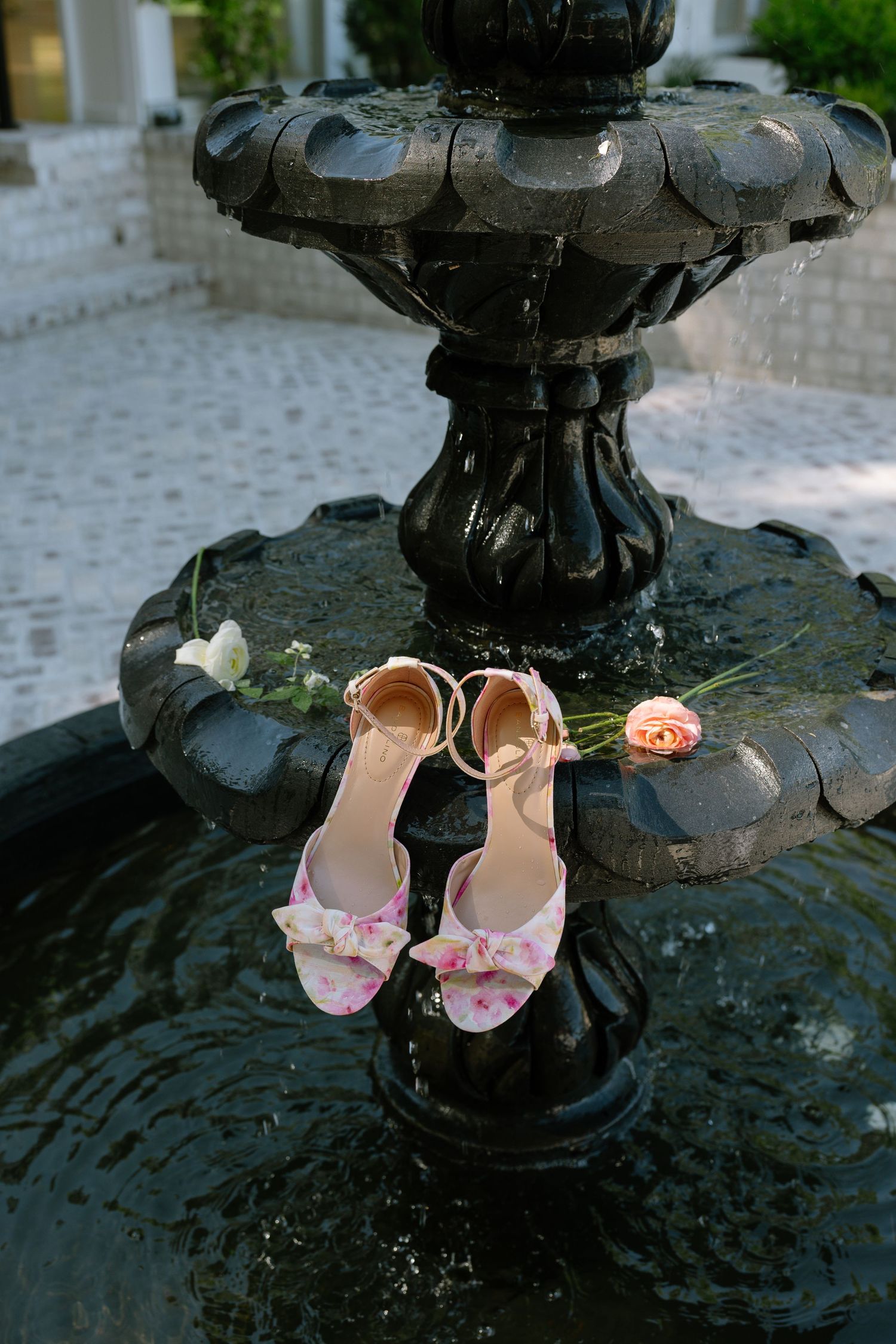 Floral bridal heels with bow detail resting on a black decorative fountain with pink roses nearby.