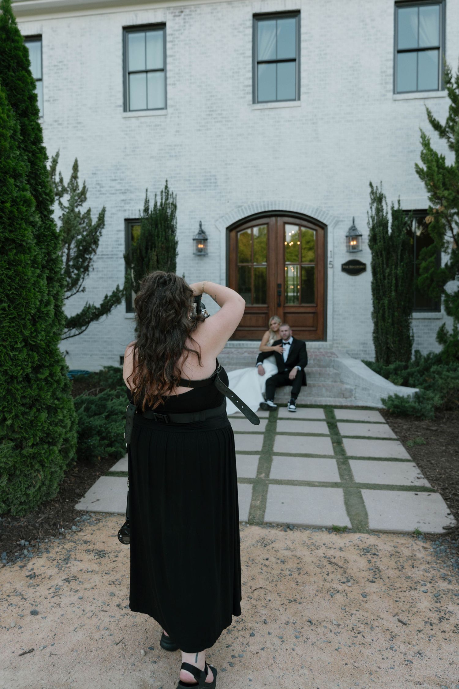 Photographer capturing bride and groom sitting on steps of white brick building with arched wooden doors.