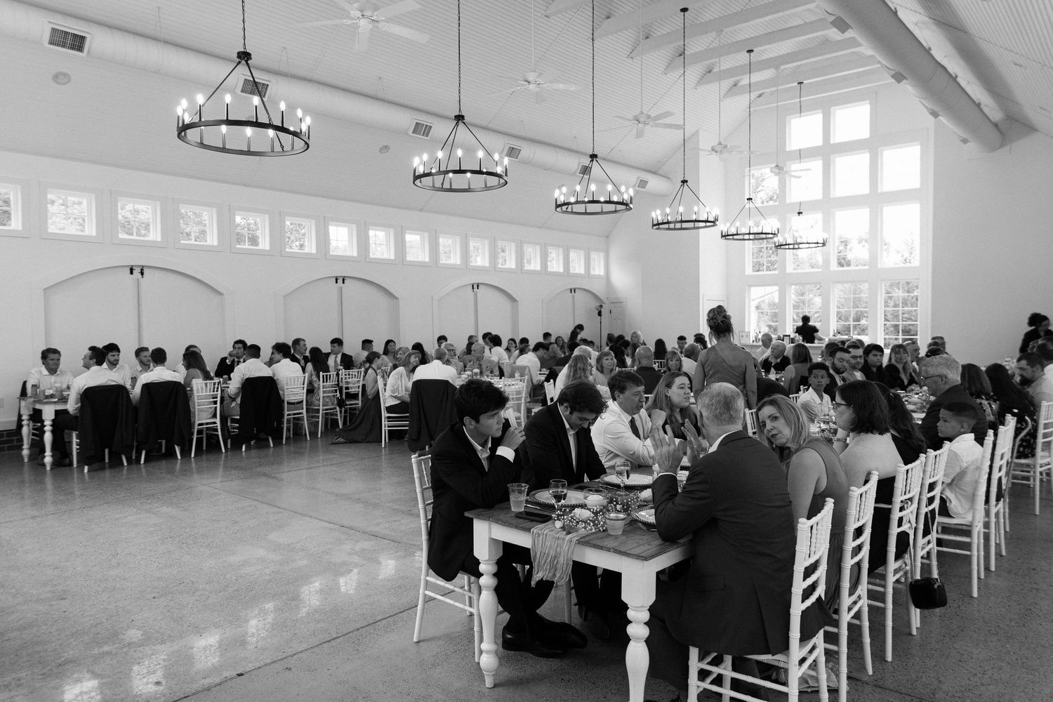 Elegant wedding reception in a bright white ballroom with chandeliers and guests dining at decorated tables.