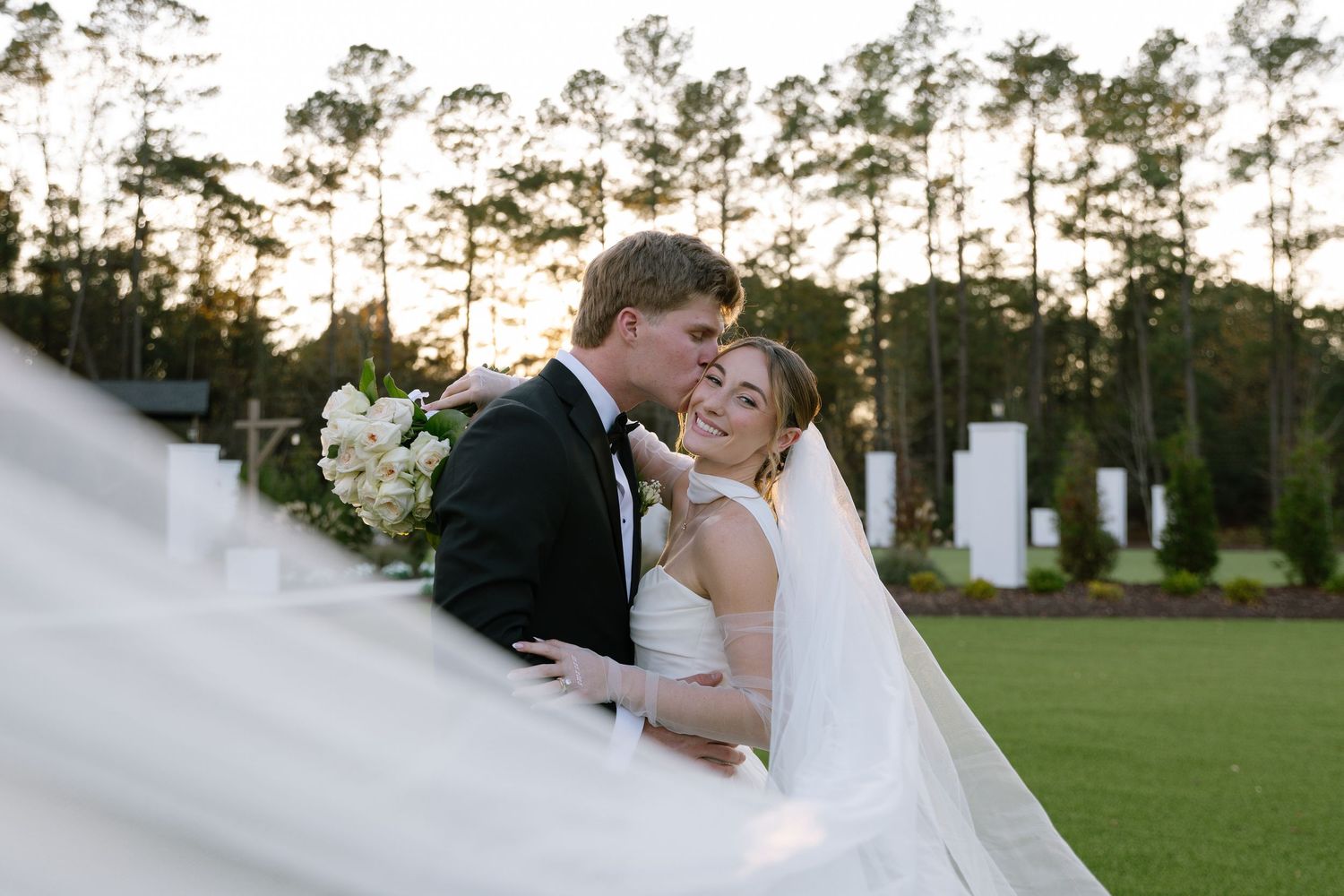 Bride and groom share romantic kiss outdoors at golf course wedding venue surrounded by tall pine trees at golden hour.