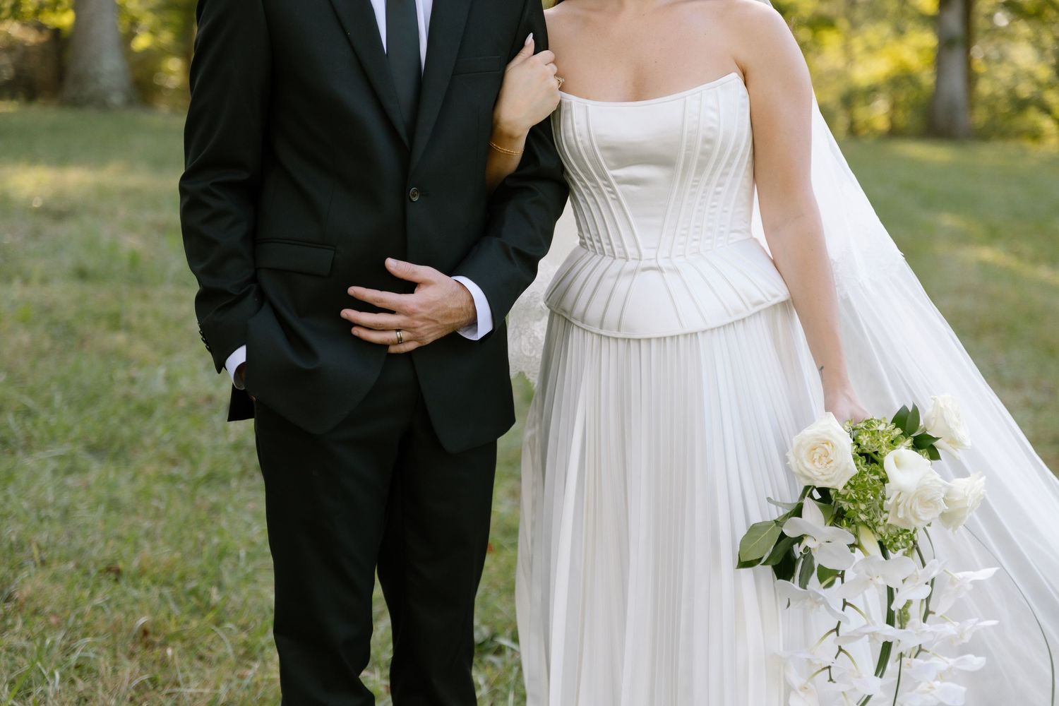 Bride in strapless white gown holding bouquet stands beside groom in black suit outdoors.