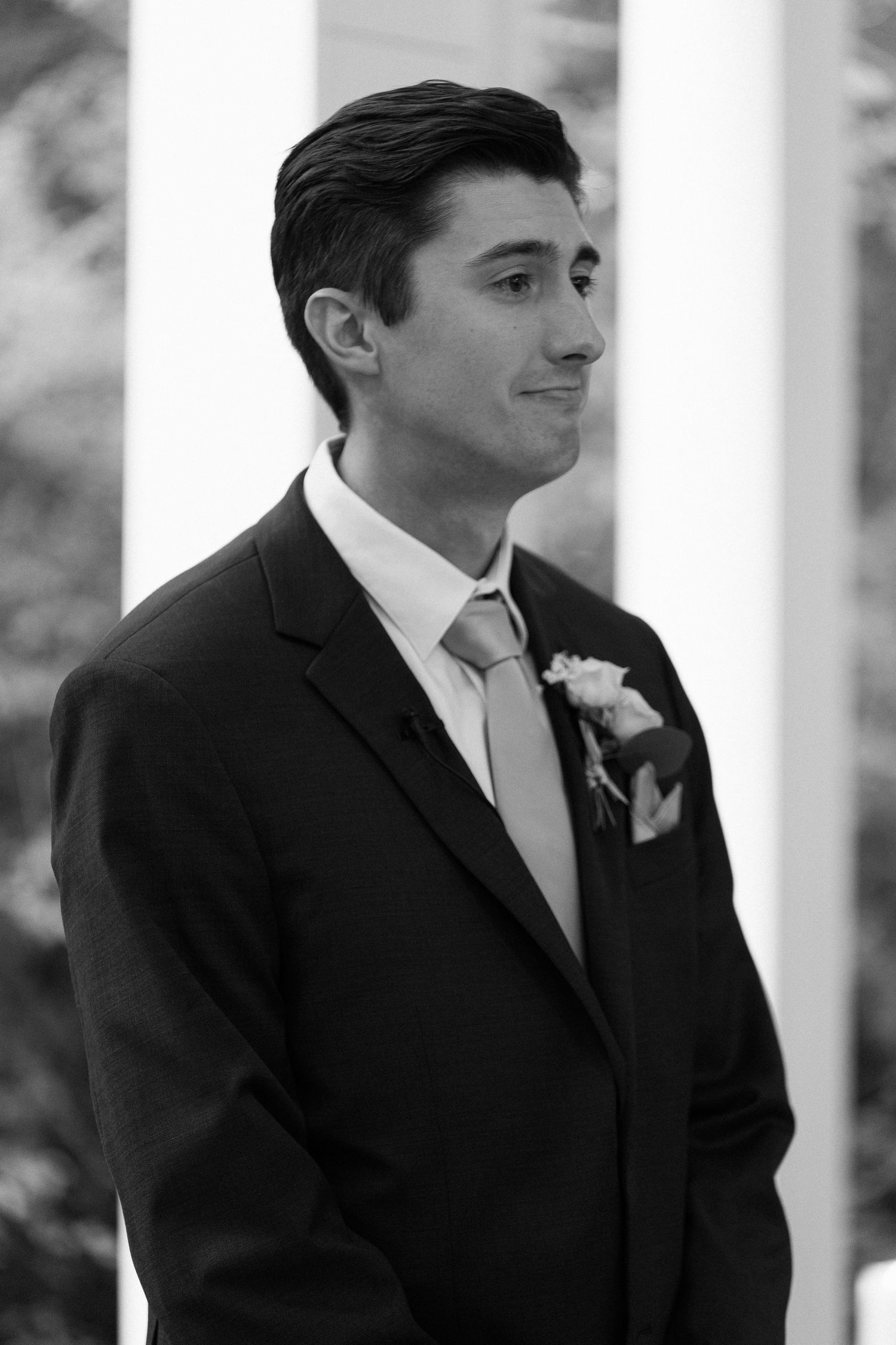Emotional groom in dark suit and boutonniere waiting at the altar during wedding ceremony, black and white.